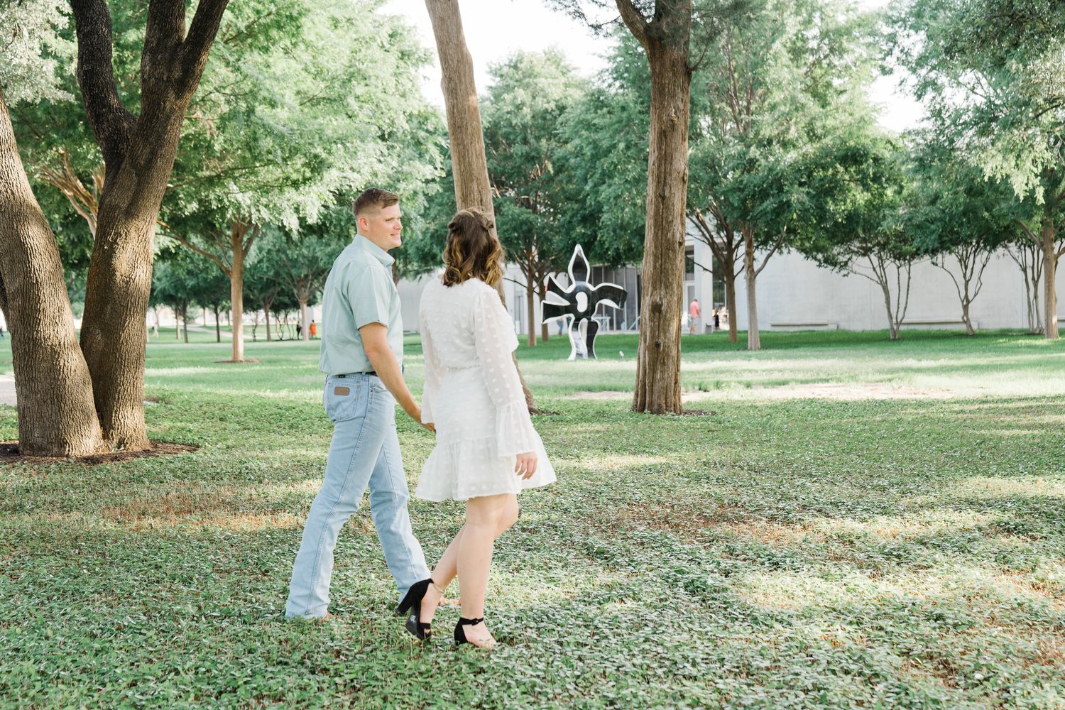 A couple walks together through a serene park setting with trees casting dappled shadows on the ground.