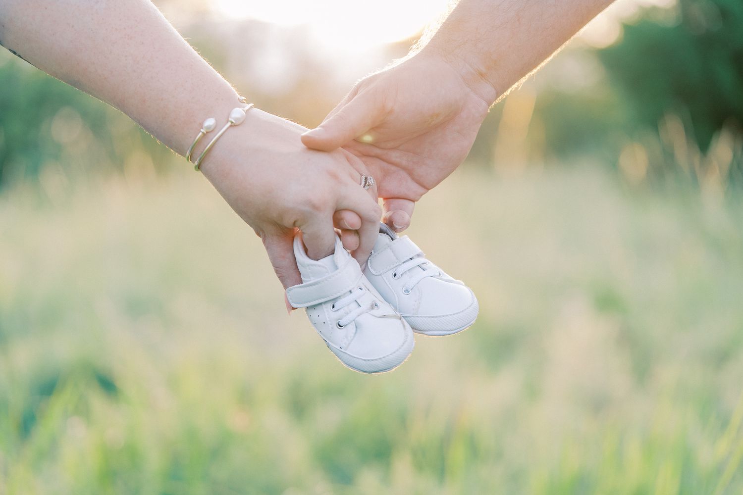 Hands hold tiny white baby shoes against blurred natural background.