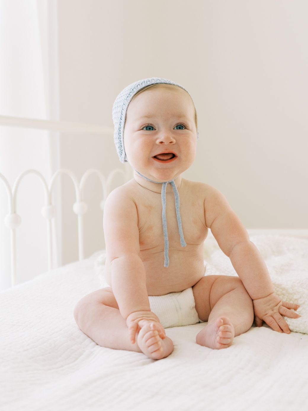 Baby wearing light blue bonnet sitting on white bedding with joyful expression.