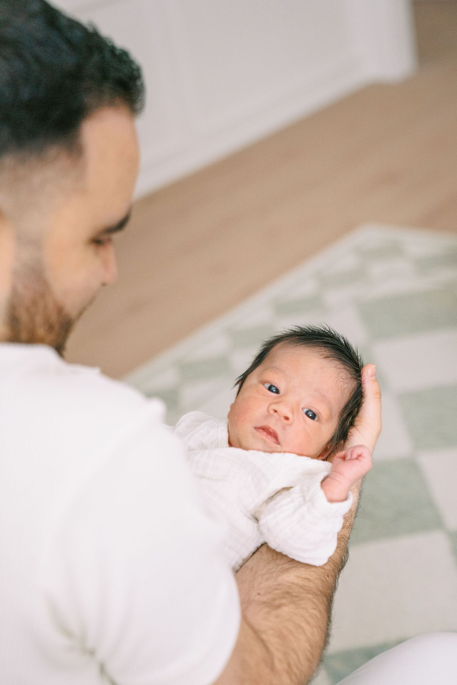 Father in white shirt cradling newborn baby with dark hair in bright home setting.