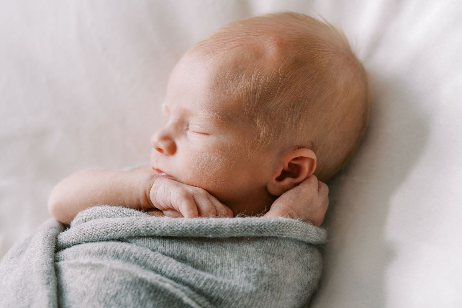 Peaceful newborn baby sleeping wrapped in a soft gray blanket, eyes closed, tiny hands tucked under chin.