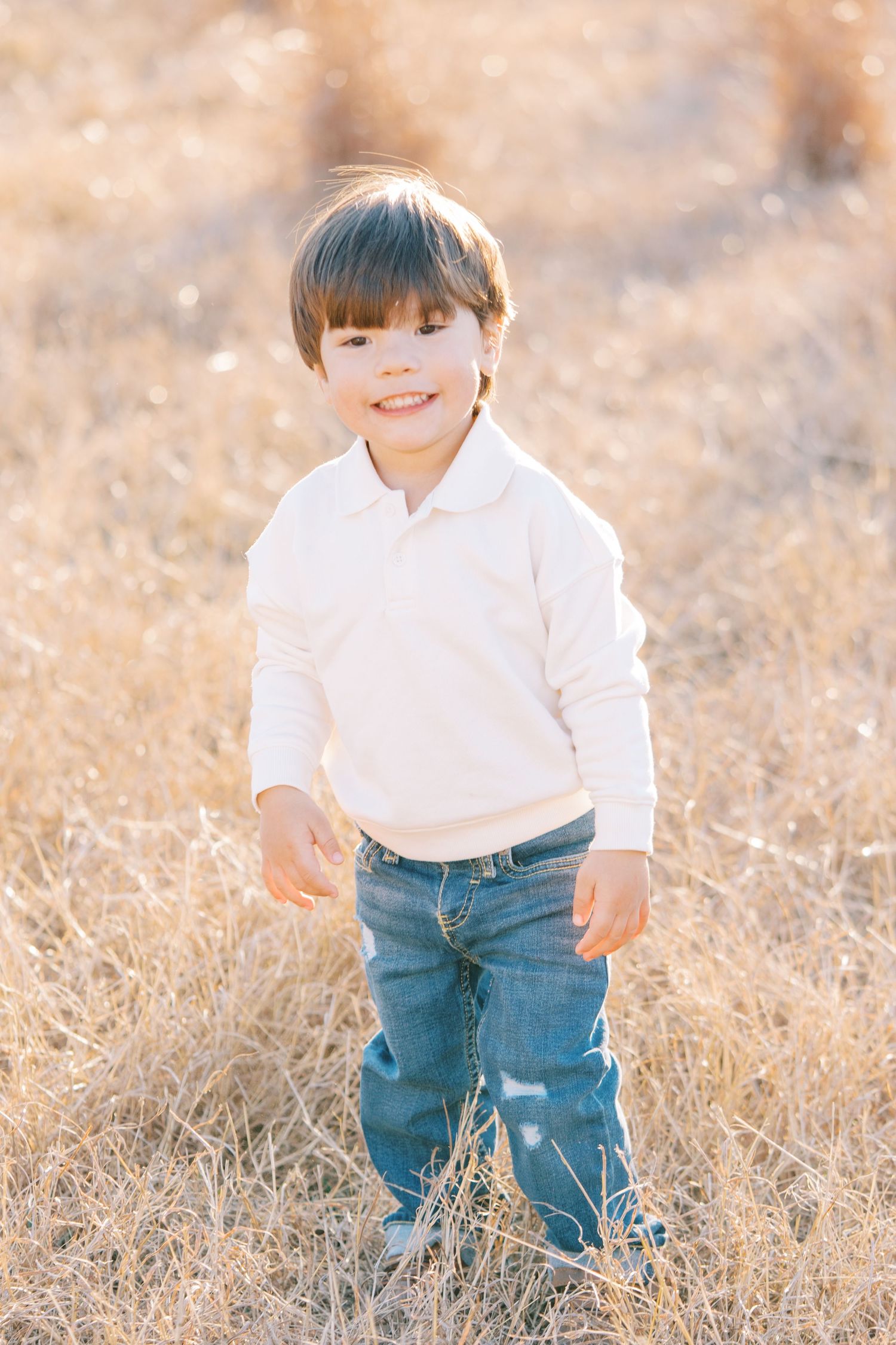 Small child in white shirt and jeans plays in golden sunlit field during outdoor photoshoot.