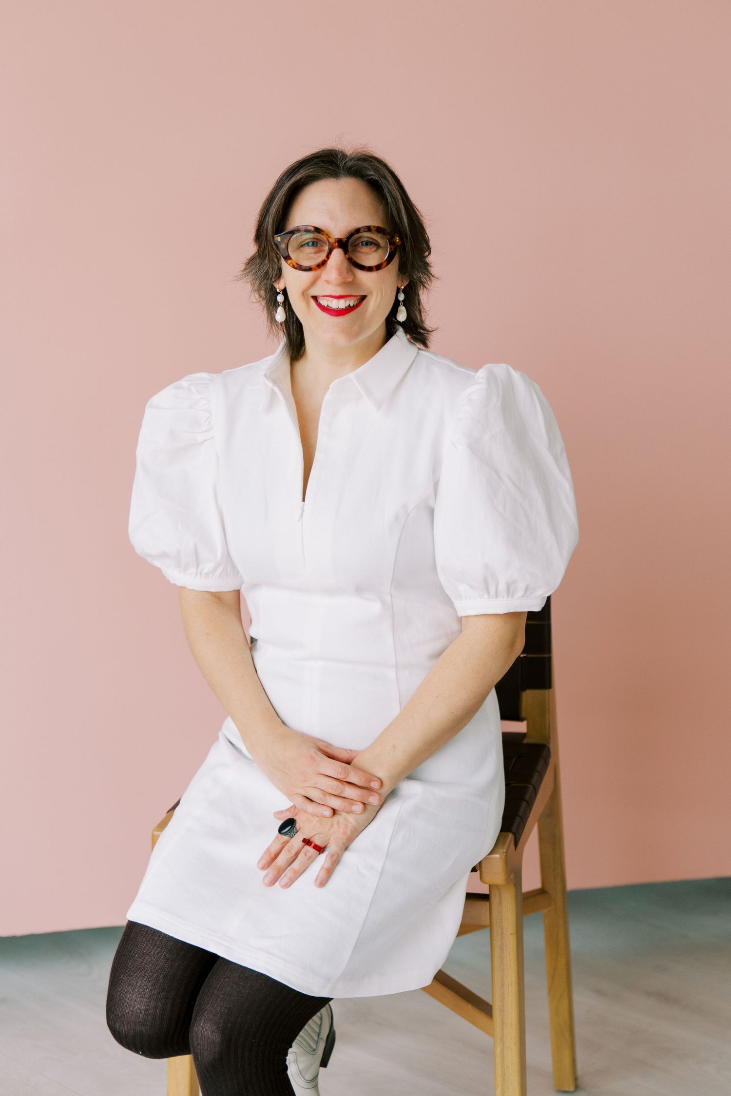 Stylish woman in white mini dress with puff sleeves sits on wooden chair against pink wall.