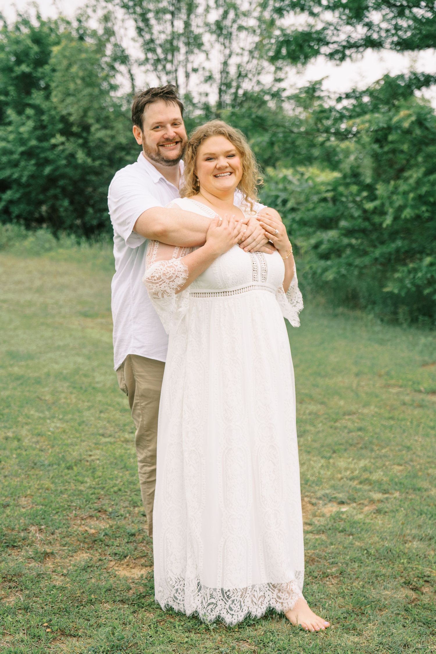 Happy couple embracing outdoors, man in white shirt hugging woman in elegant white lace dress on green lawn.