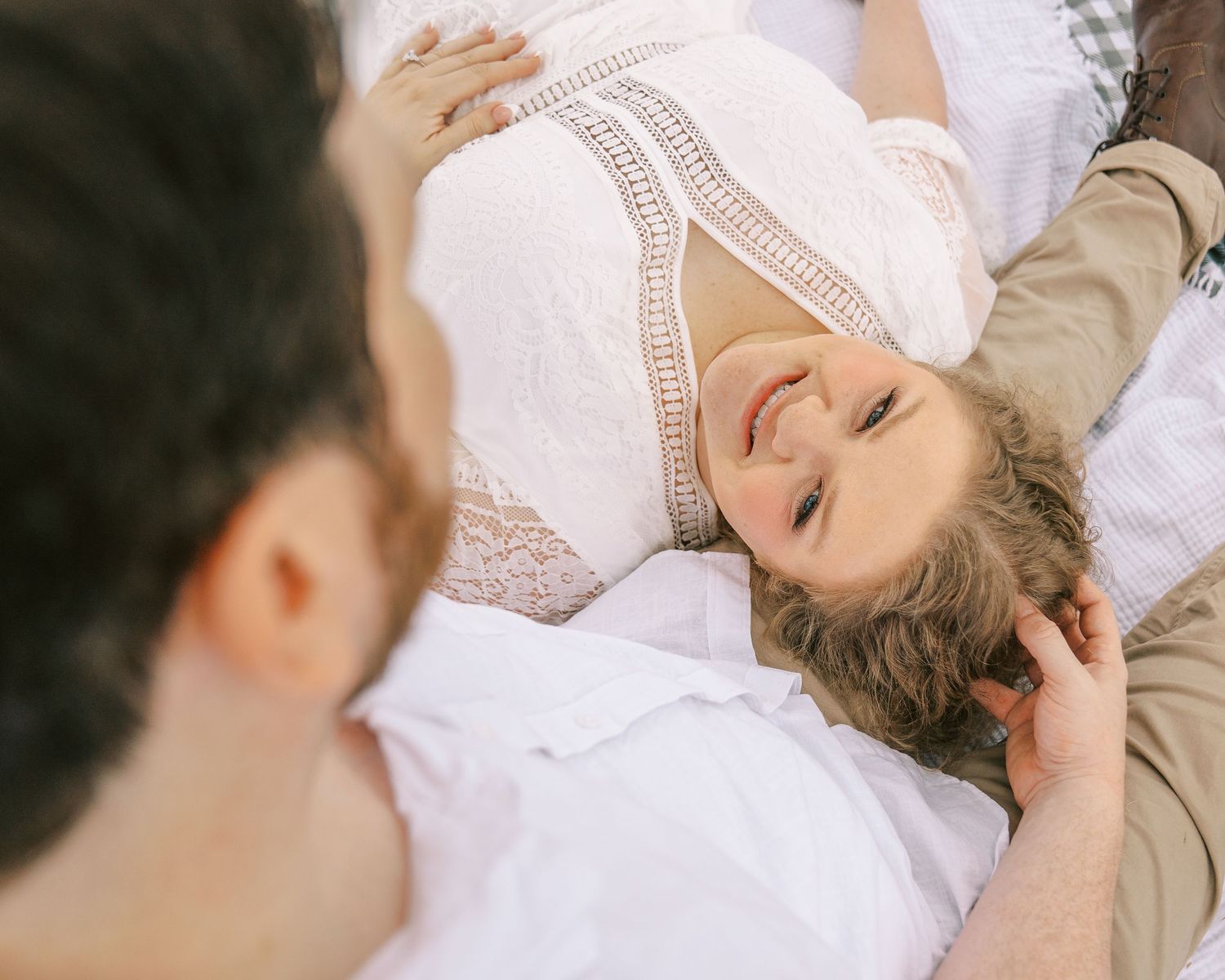 Smiling woman in white lace dress lying down looking up at her partner during outdoor couples photography.