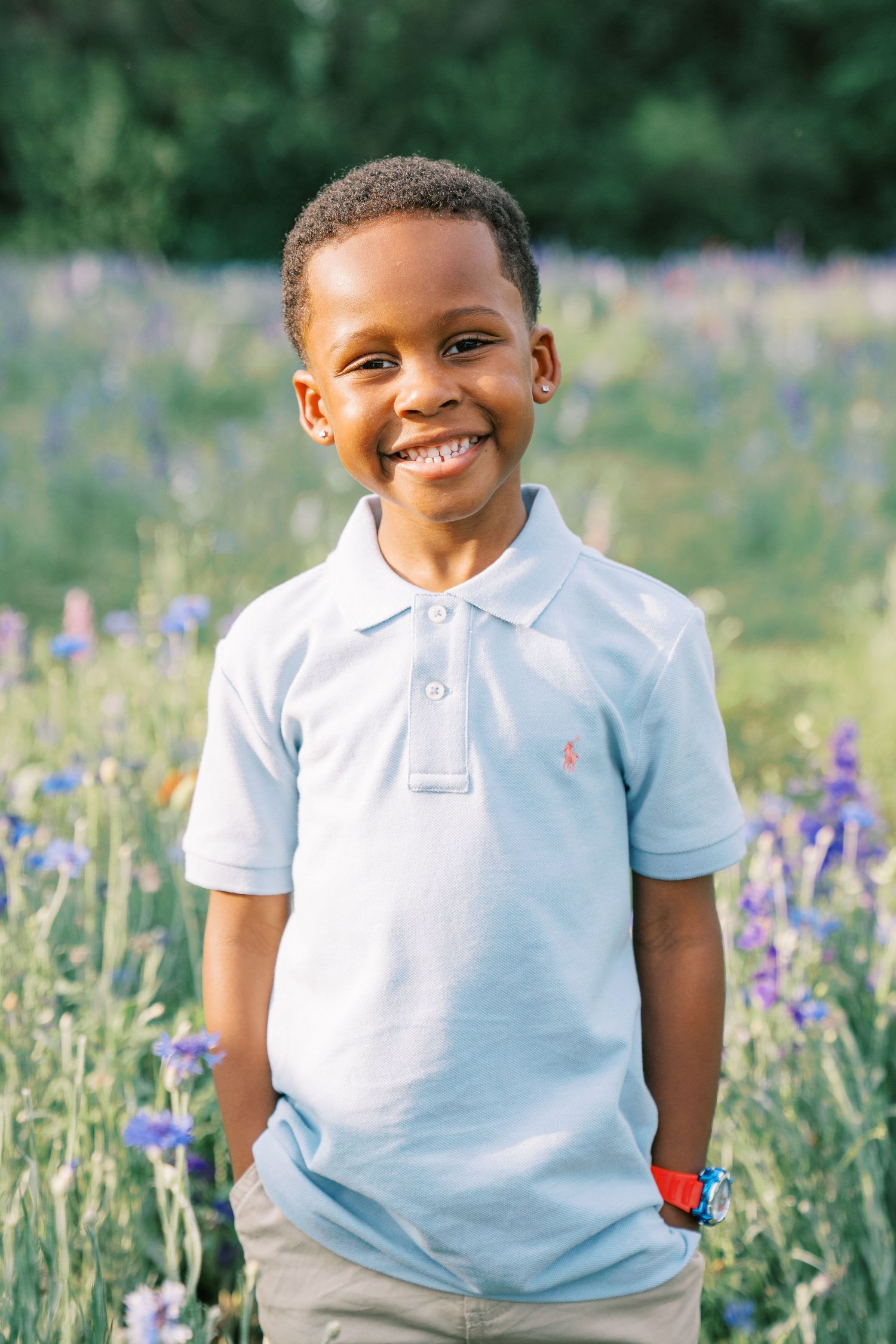 Smiling young boy in a light blue polo shirt standing in a colorful wildflower field outdoors.