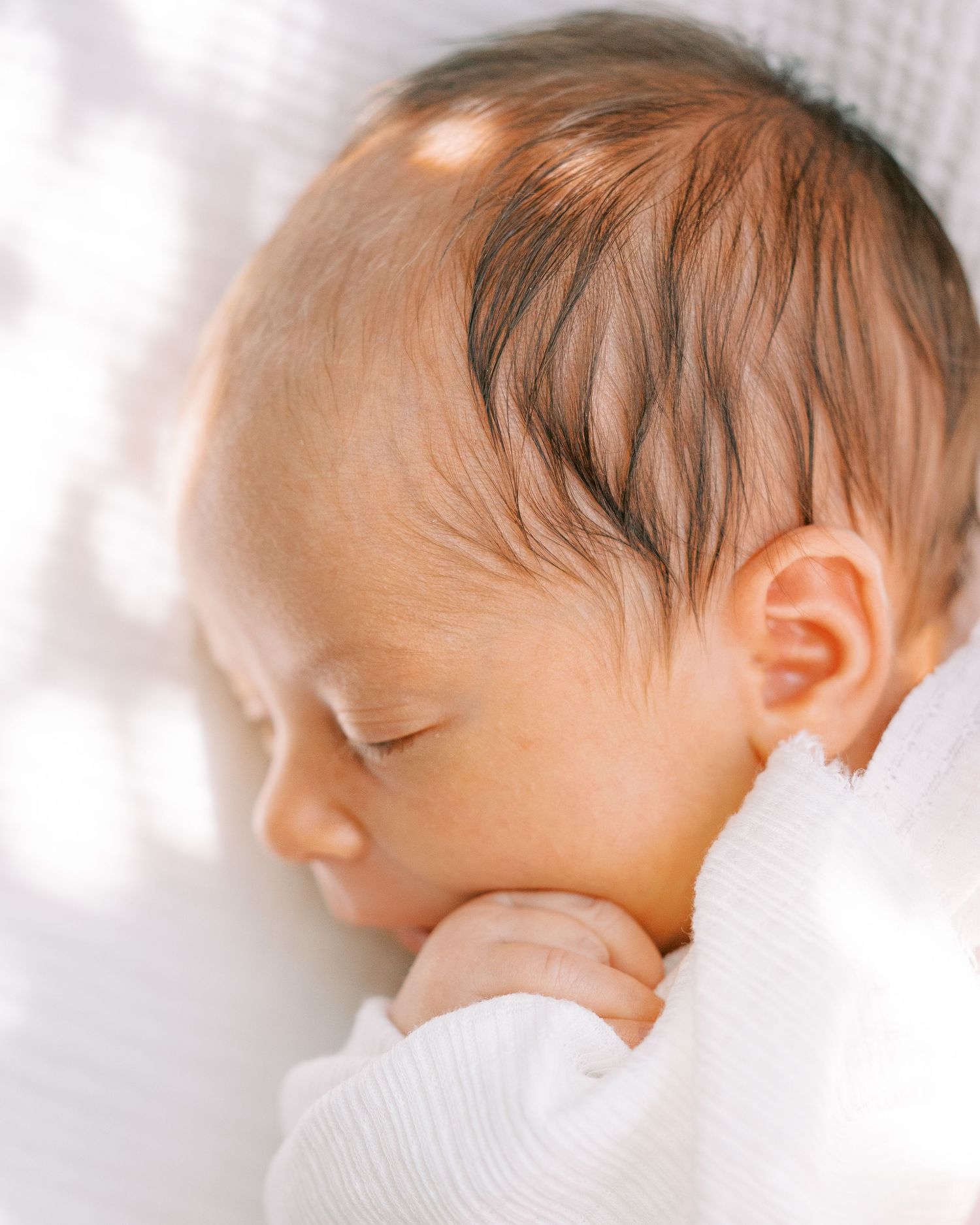 Sleeping baby in white clothing rests peacefully against soft white bedding in gentle natural light.