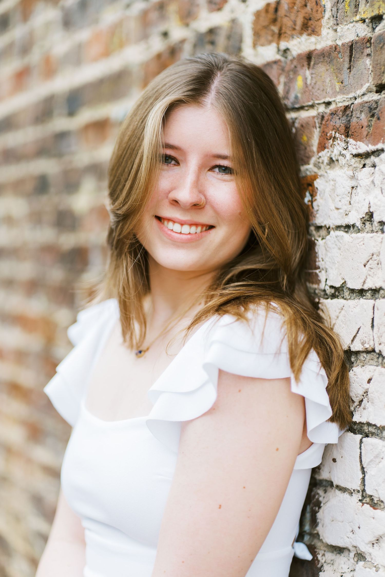 A person in a white ruffled top poses against a rustic brick wall with natural lighting.