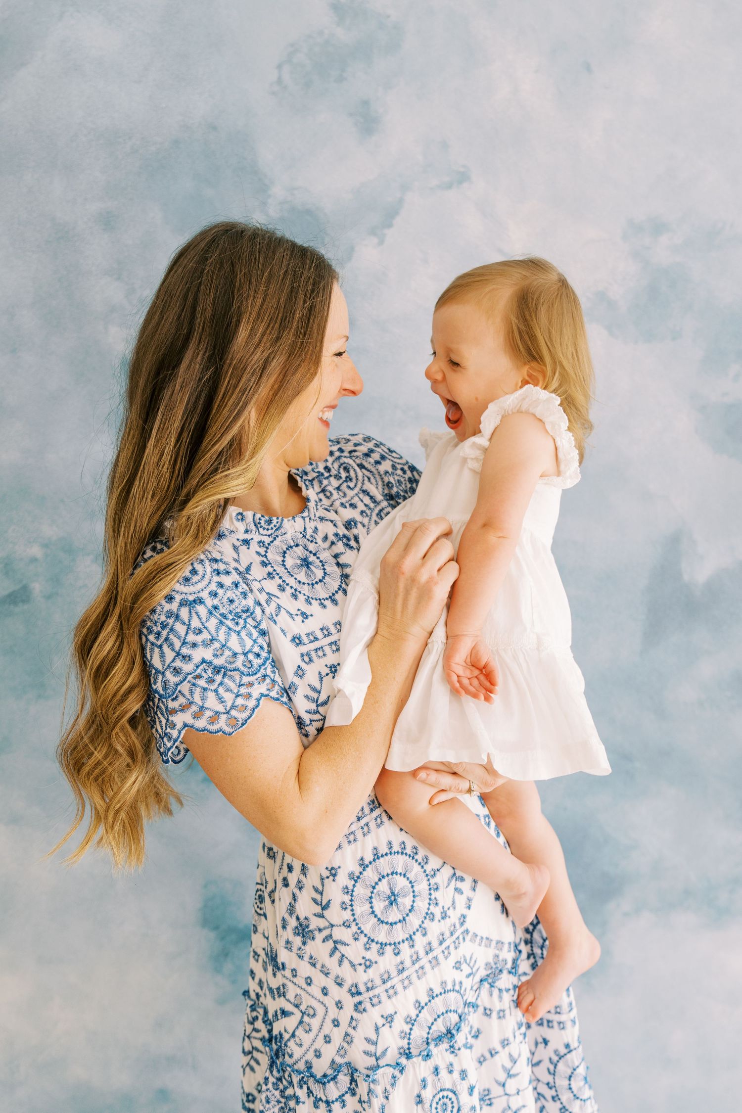 Mother and baby in matching blue and white dresses share joyful moments against cloudy backdrop.