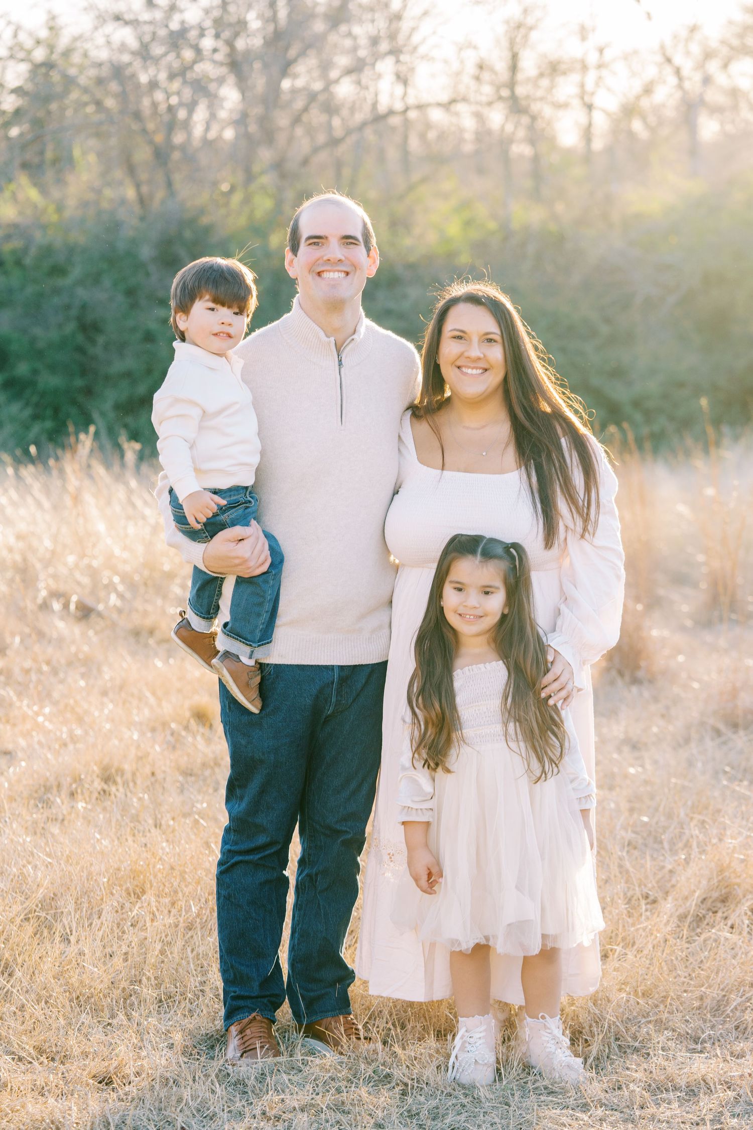 Family wearing coordinated white and blue outfits poses together in a sunlit field of tall grass at golden hour.