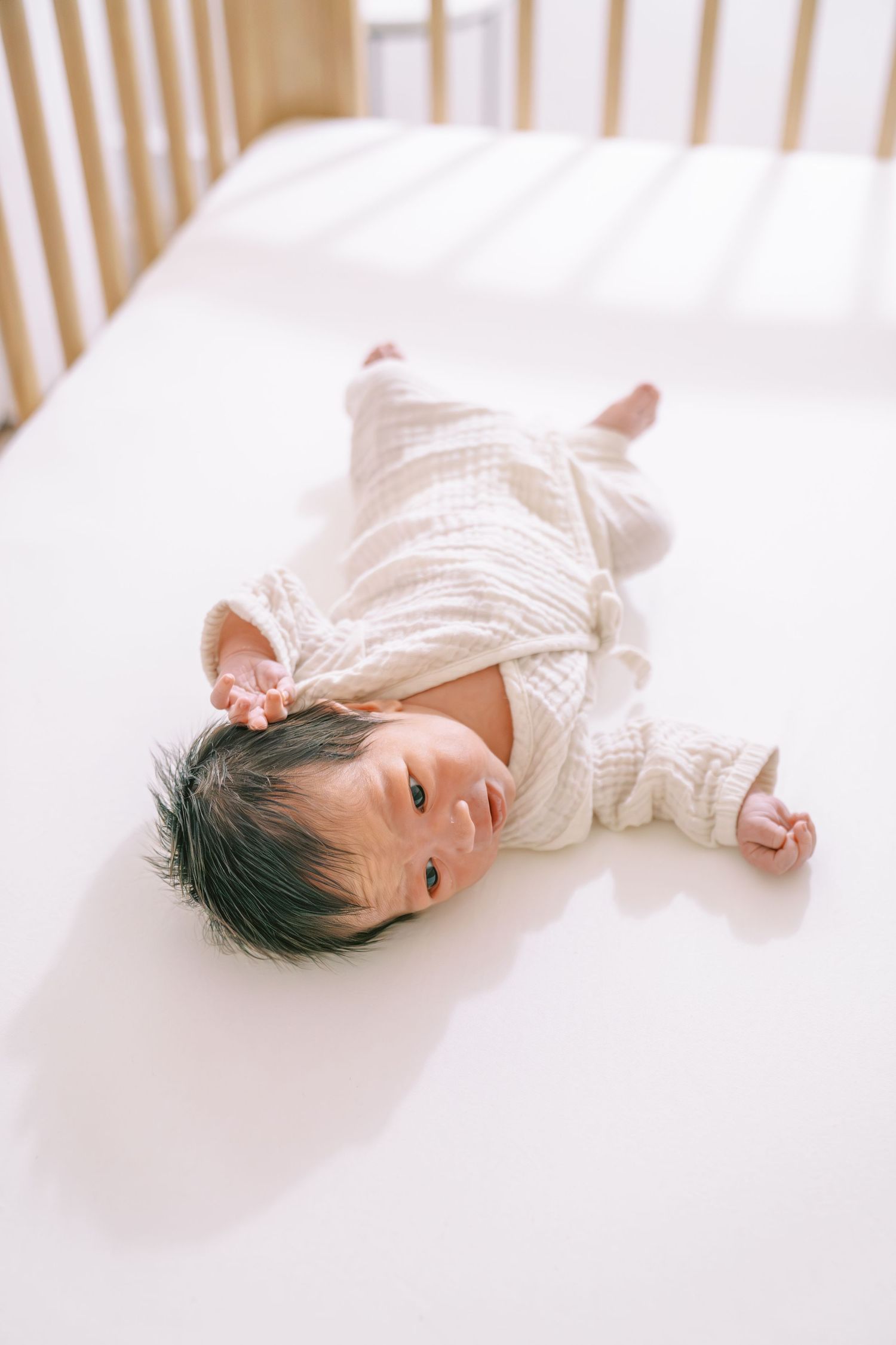 Newborn baby in white knit outfit lying on back in a white crib, looking up with dark hair and tiny hands.