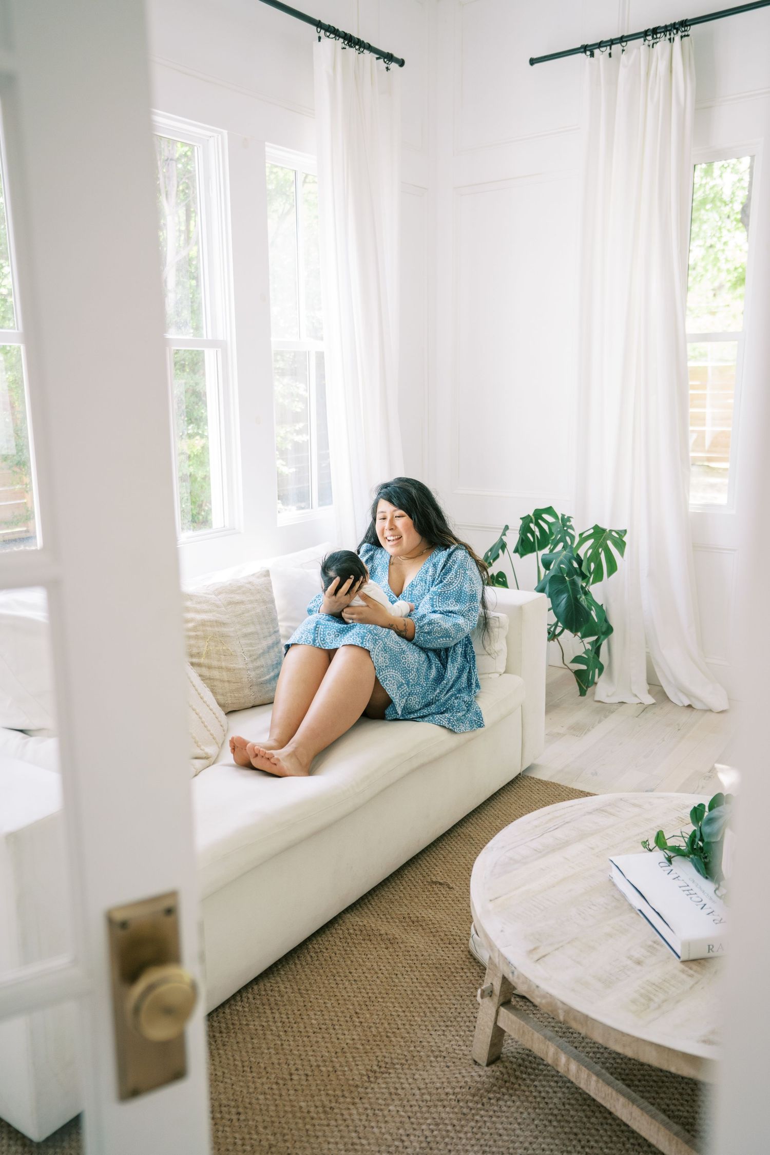 Woman in blue floral dress relaxing on white sofa using smartphone in bright, airy living room with monstera plant.
