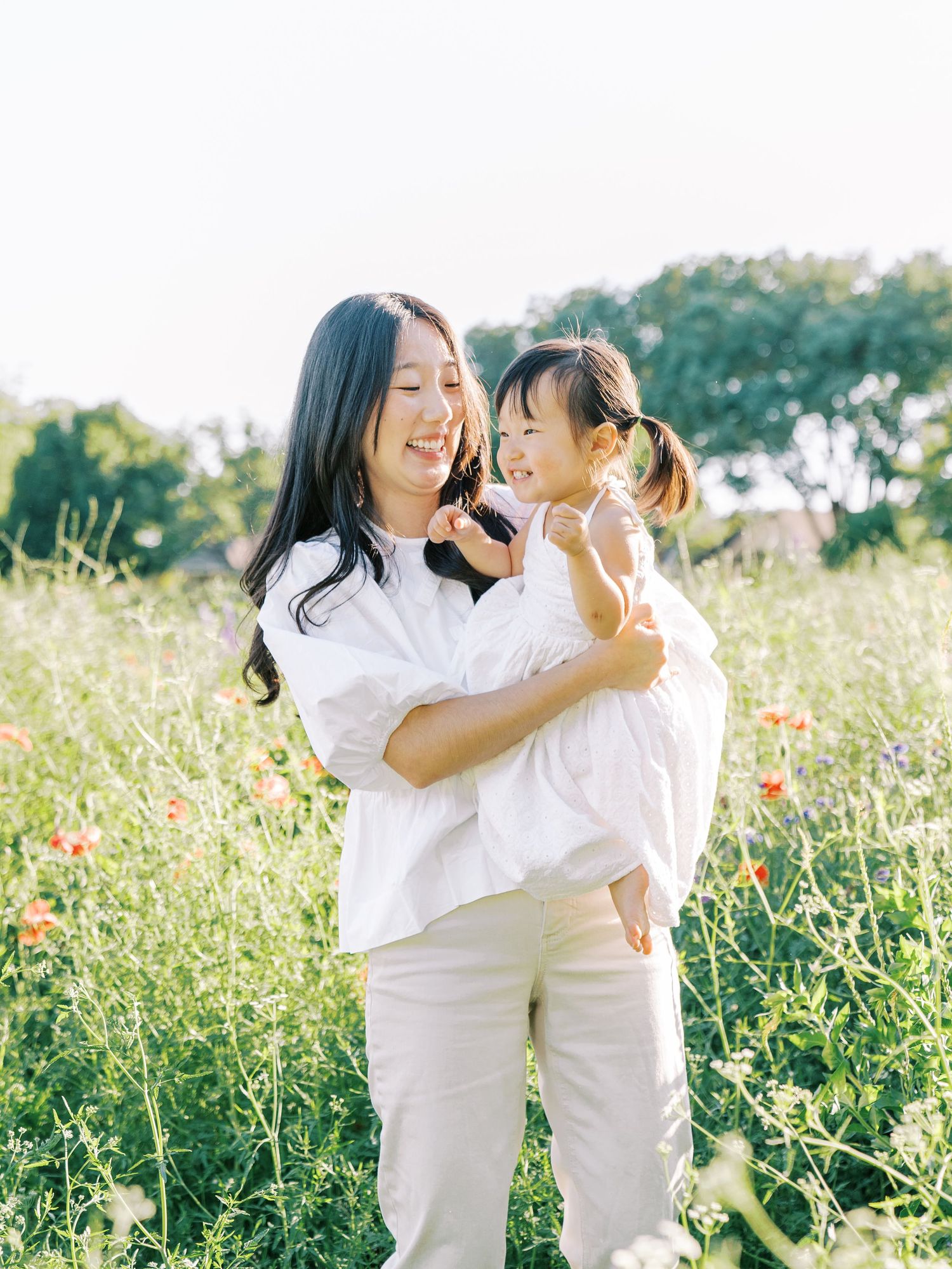Two people in white outfits share tender moments in a sunlit wildflower meadow during a photo session.