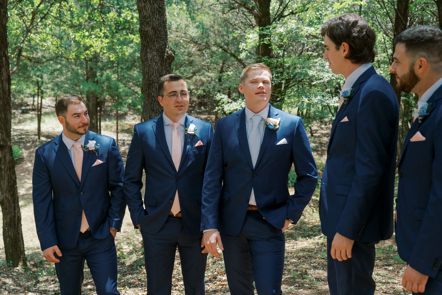 A group in matching navy blue suits and blush pink ties stand together in a wooded outdoor setting.