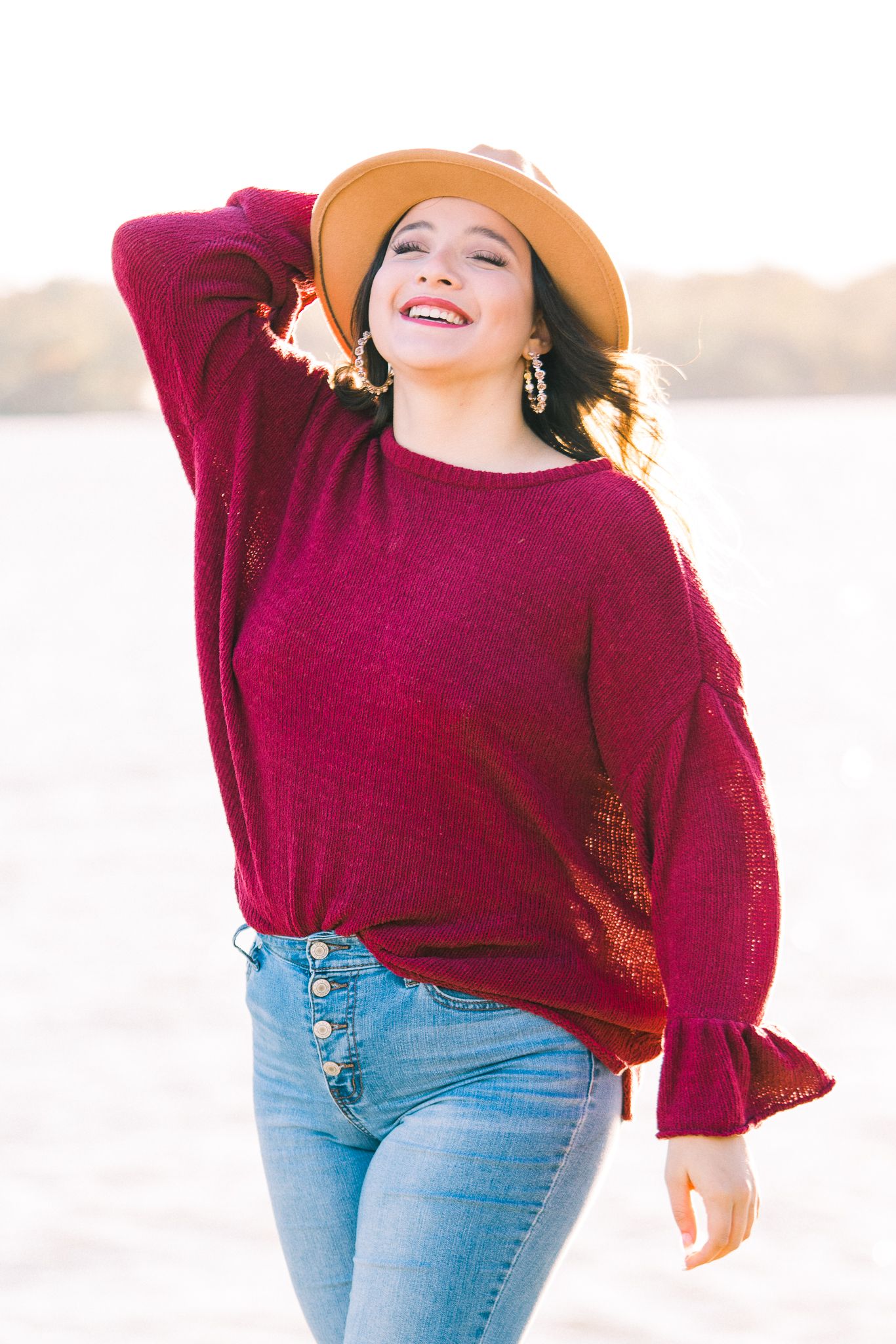 Someone wearing a cozy maroon sweater and blue jeans stands against a bright waterfront backdrop.