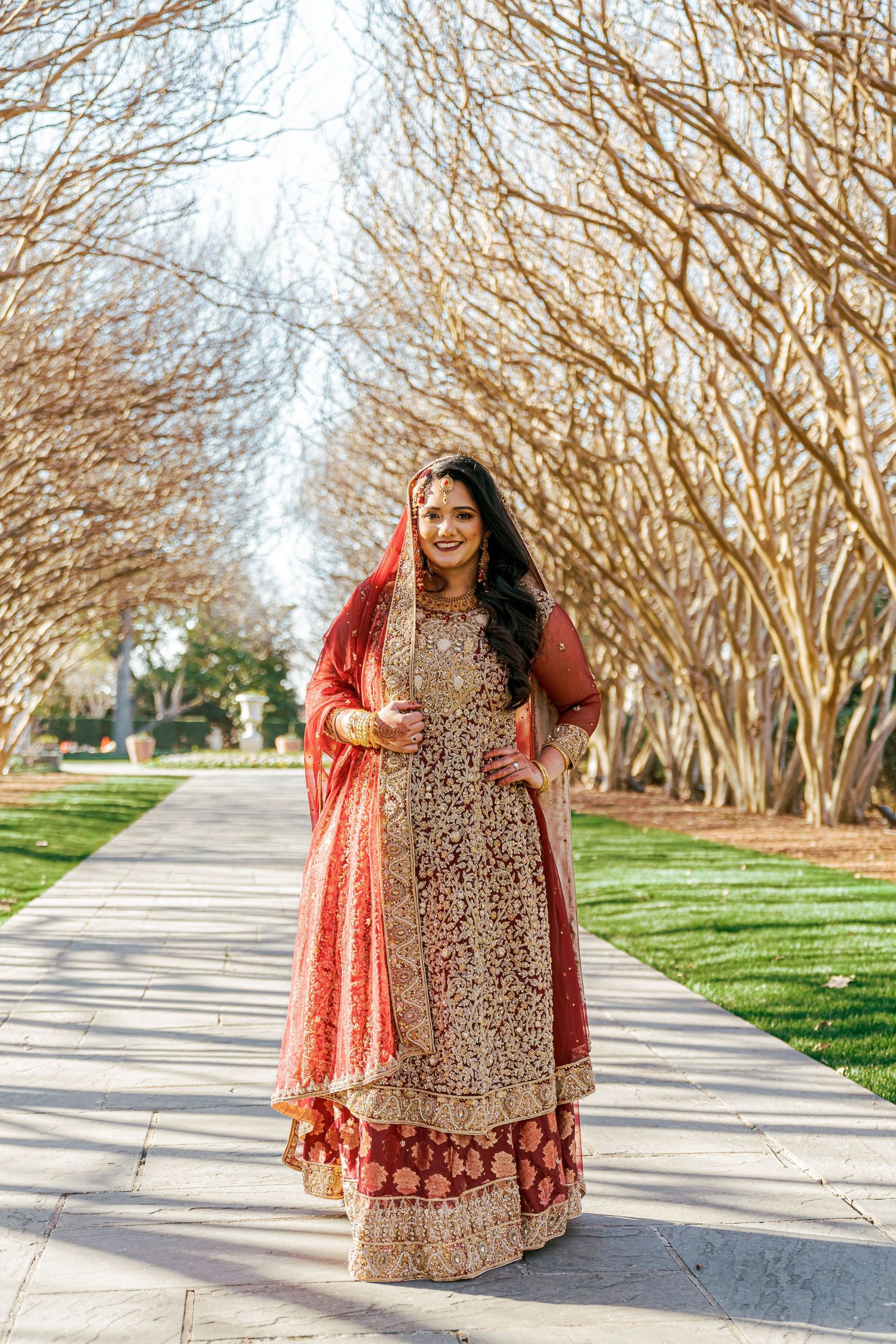 Bride in red and gold lehenga poses along tree-lined pathway during winter wedding photoshoot.