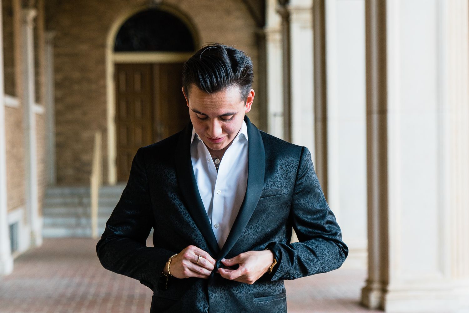 A groom in a black suit adjusts his tie while getting ready in an elegant hallway with arched windows.