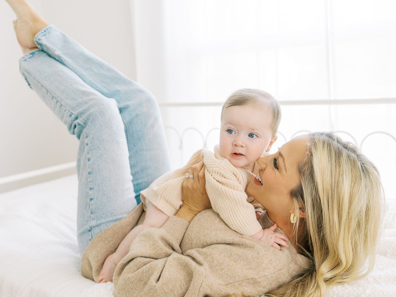Mother and baby playing and laughing together on white bedding in natural light.