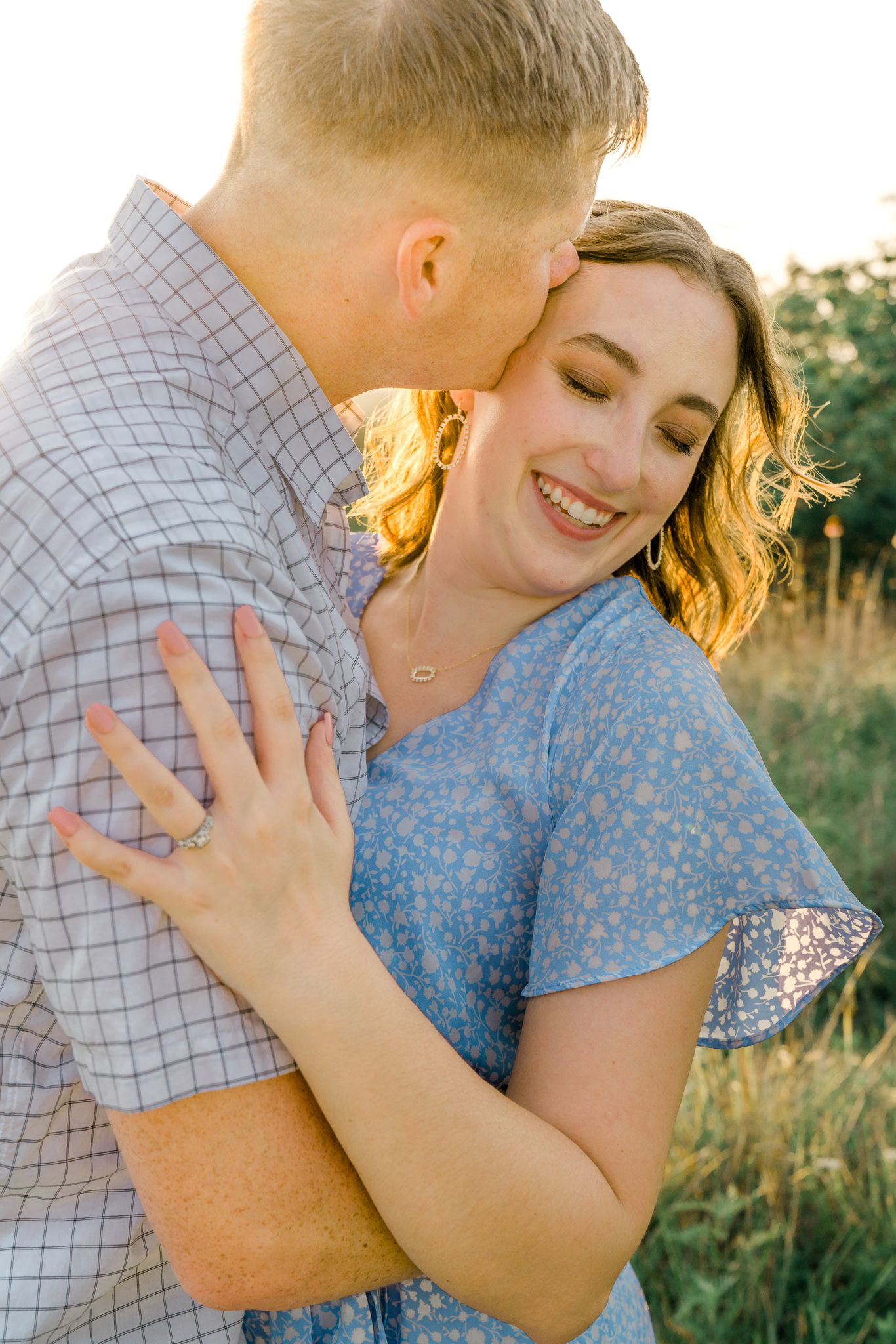 A romantic couple embraces in a field at golden hour wearing a blue floral dress and checkered shirt.