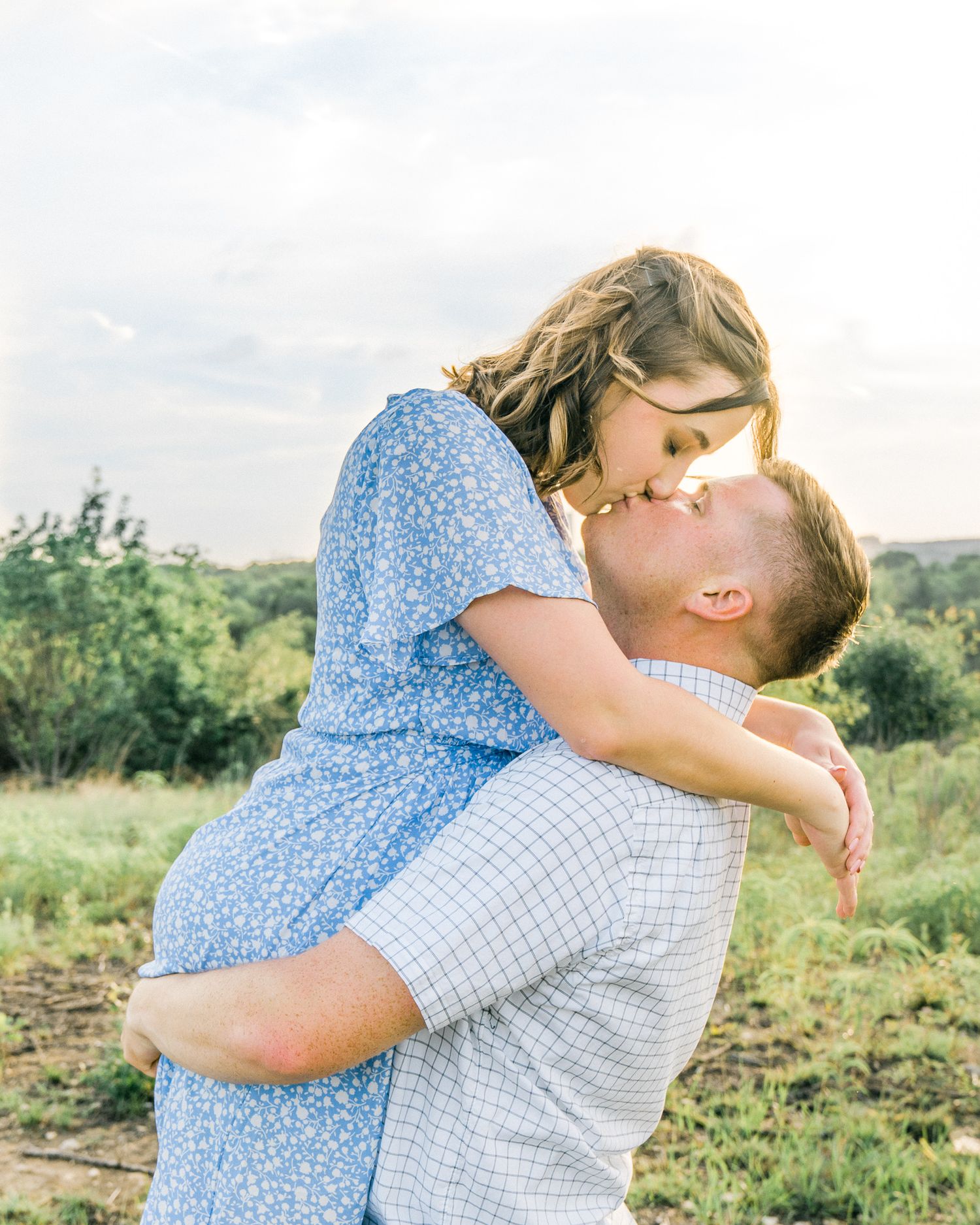 Two people share an intimate embrace in a golden field during sunset wearing blue and white clothing.