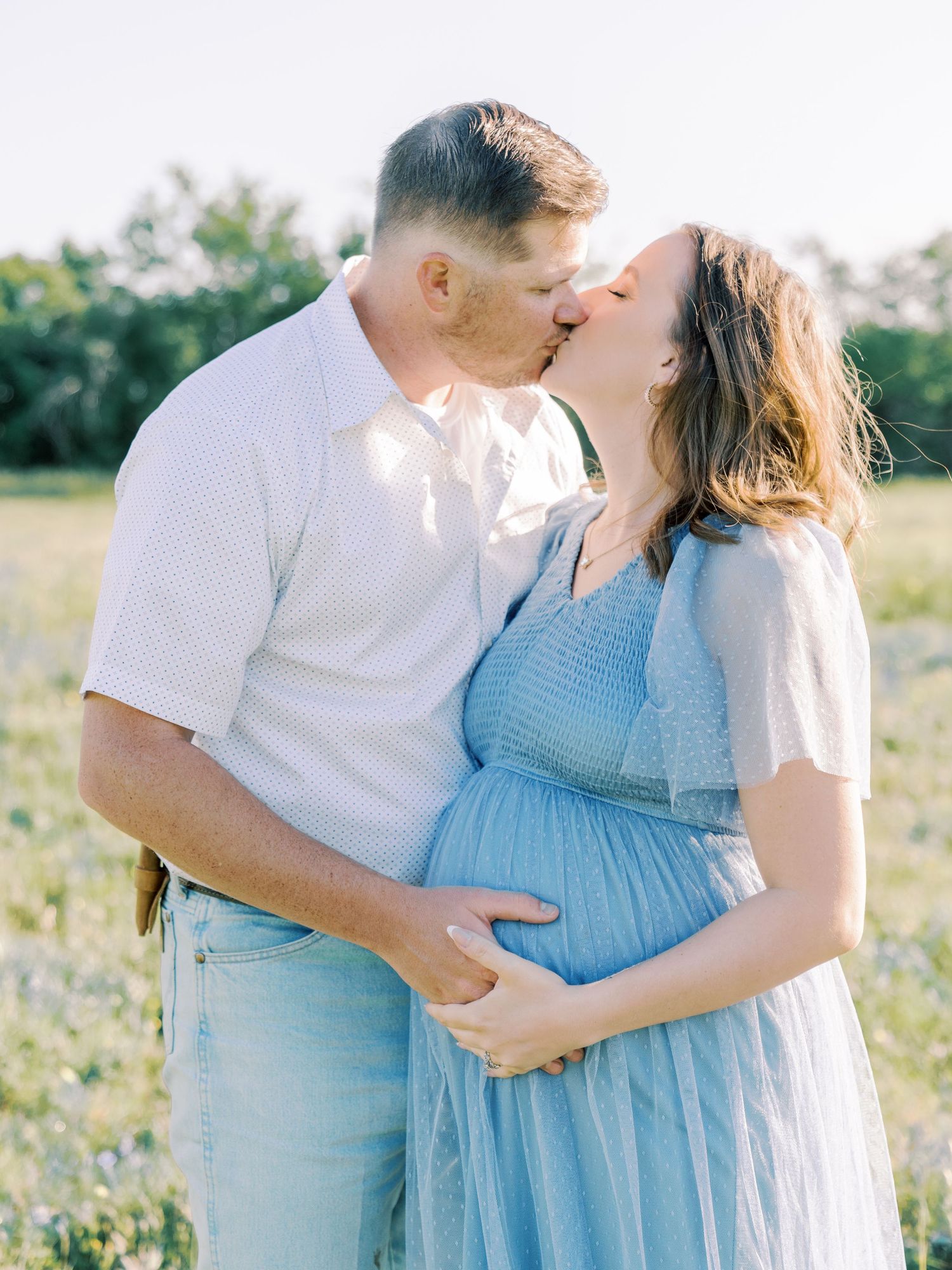 A couple embraces in a field wearing coordinating blue and white outfits during a maternity shoot.