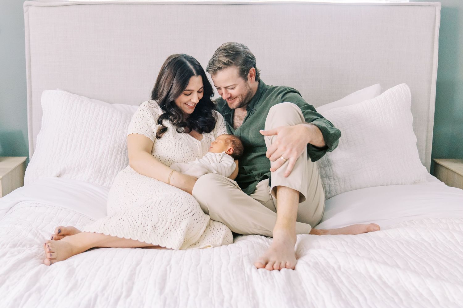 Parents cuddle with newborn baby on white bed in soft natural lighting.