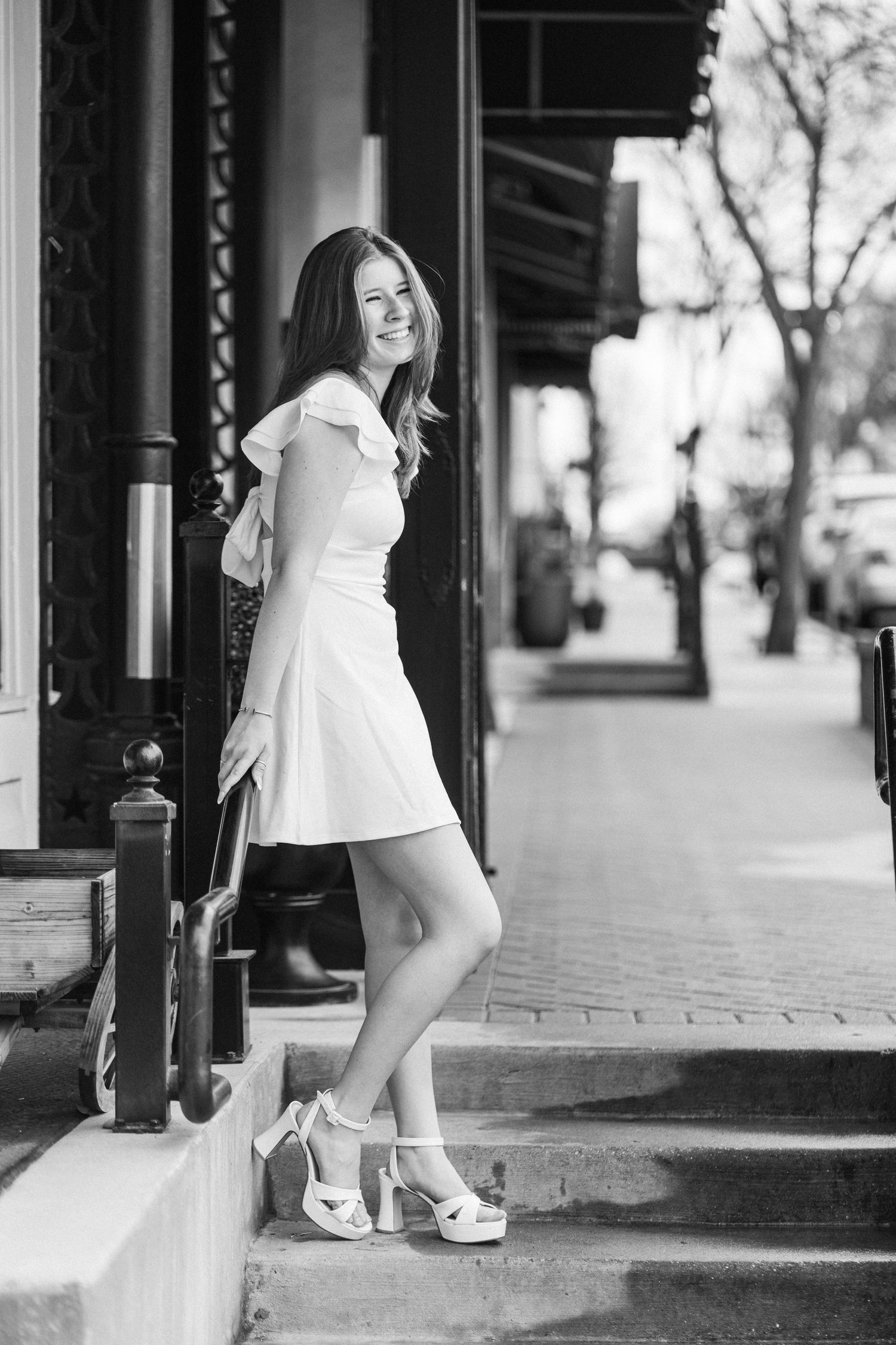Black and white portrait of someone in a white dress and heels posing on steps outside a storefront.
