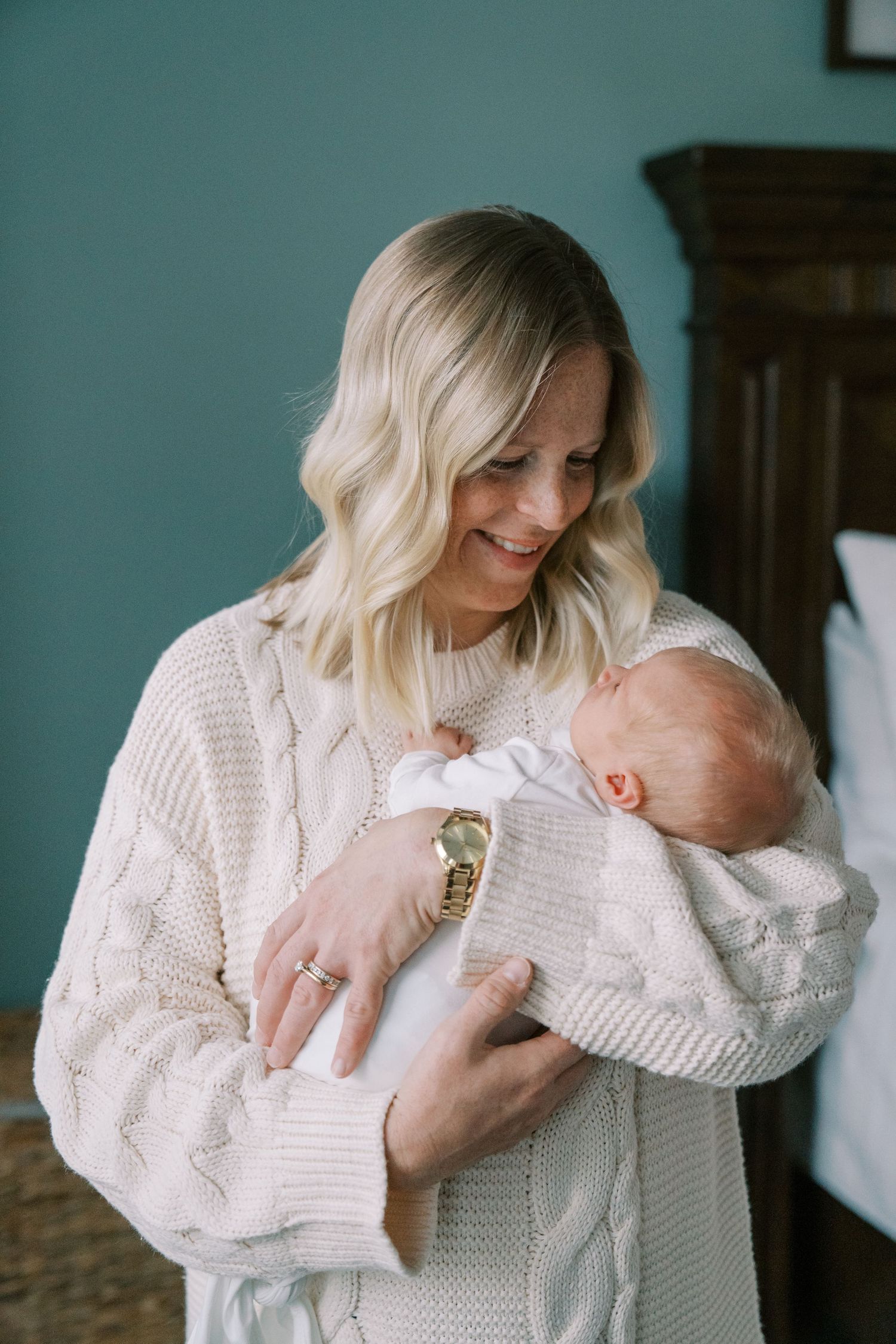 Mother in cream cable knit sweater cradles newborn baby against turquoise wall.