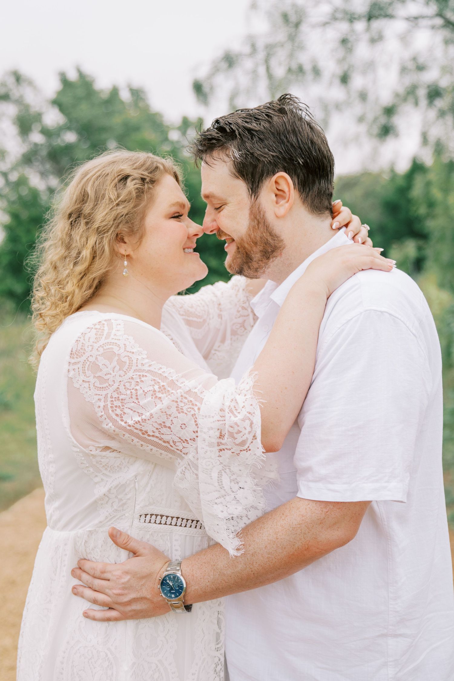 Couple in white outfits sharing romantic nose-to-nose moment outdoors during maternity photo shoot.