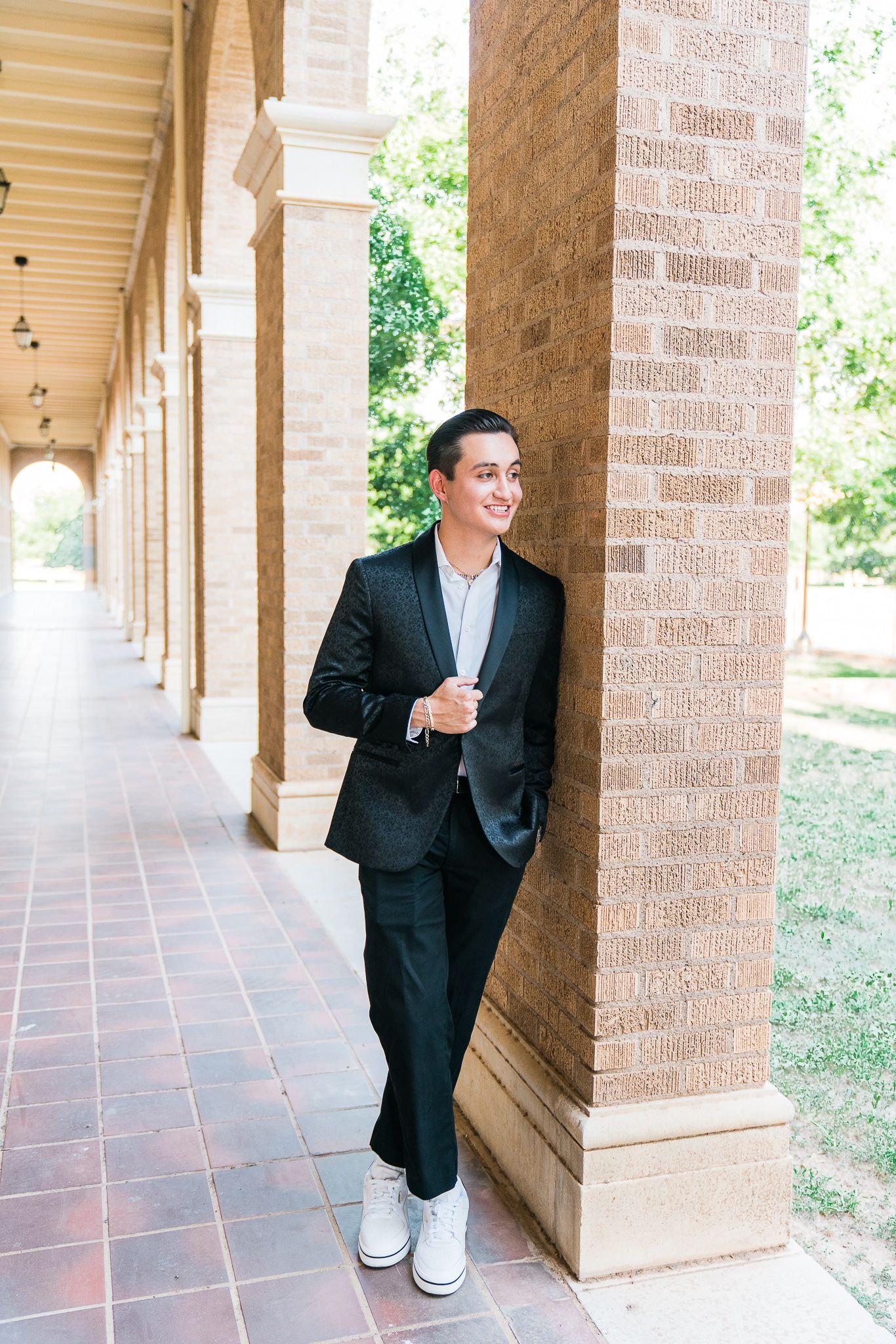 A person in a dark suit and white sneakers stands casually against a brick column in an outdoor colonnade.