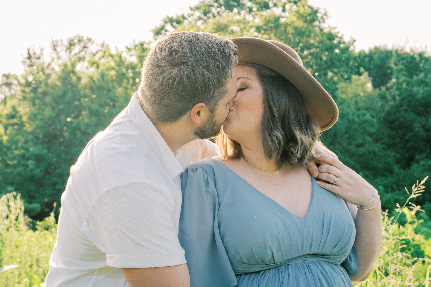 A couple shares a tender moment in a sunlit outdoor setting with lush greenery in the background.