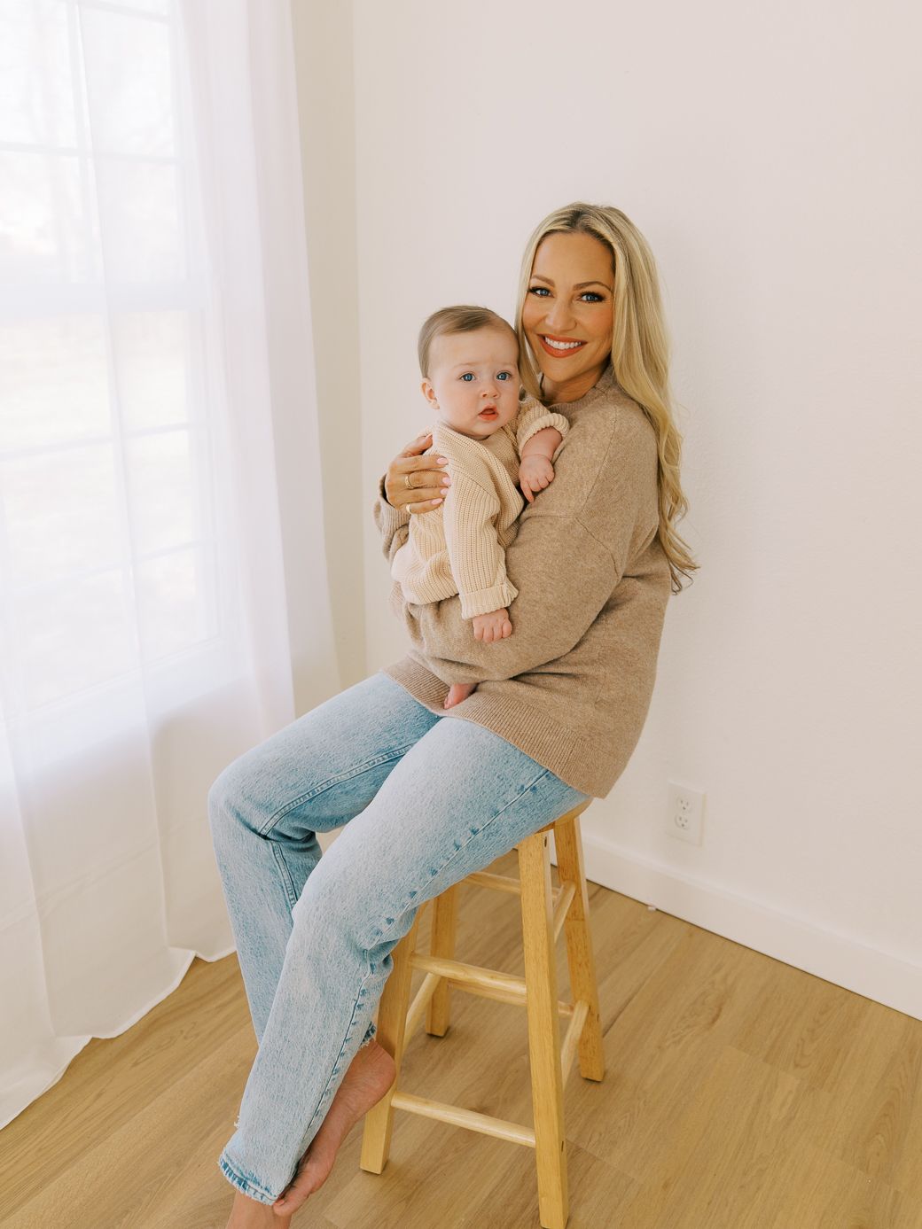Mother and baby in matching beige sweaters posing on wooden chair against white wall.