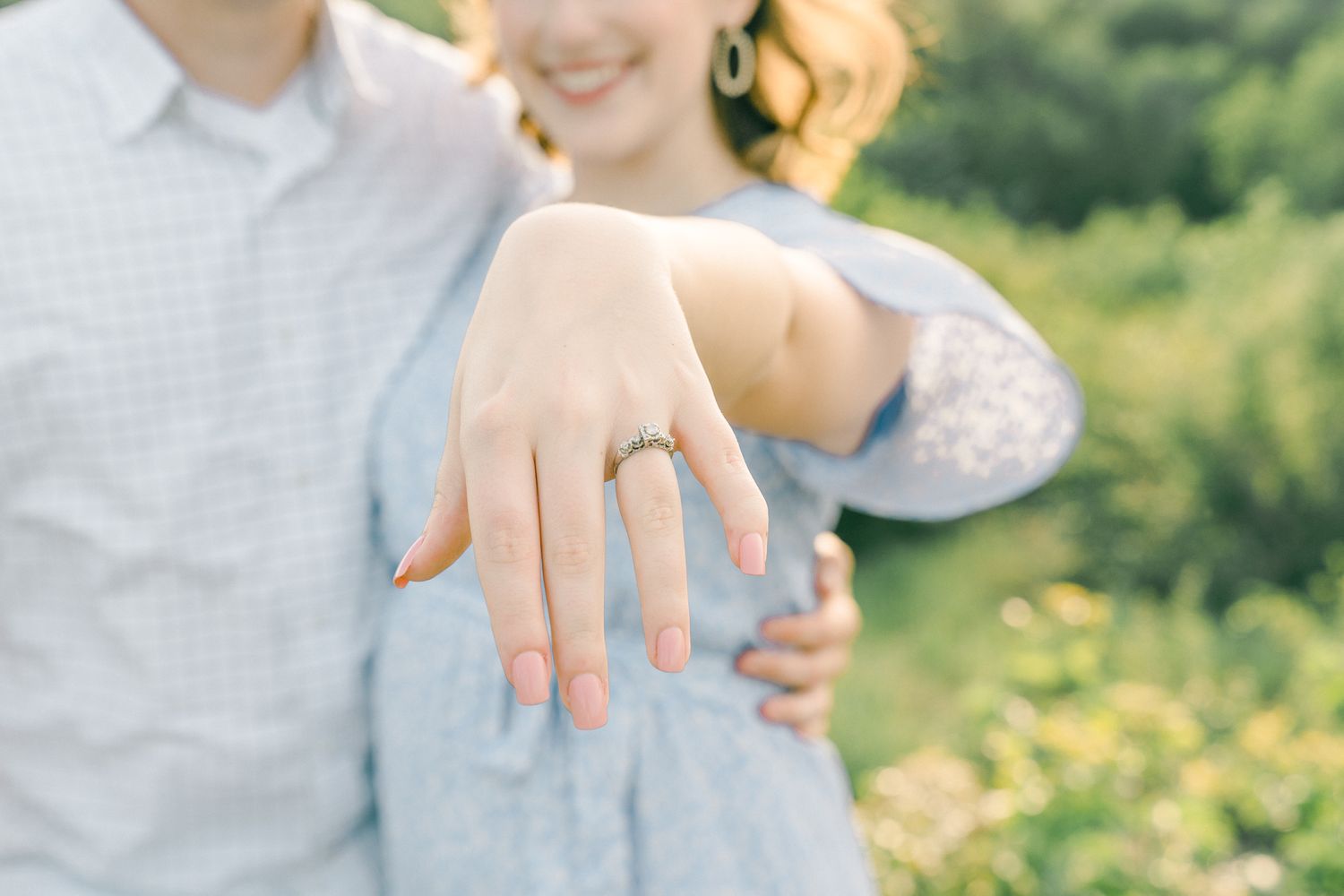 A close-up shot of a hand displaying an engagement ring against a blurred natural background.