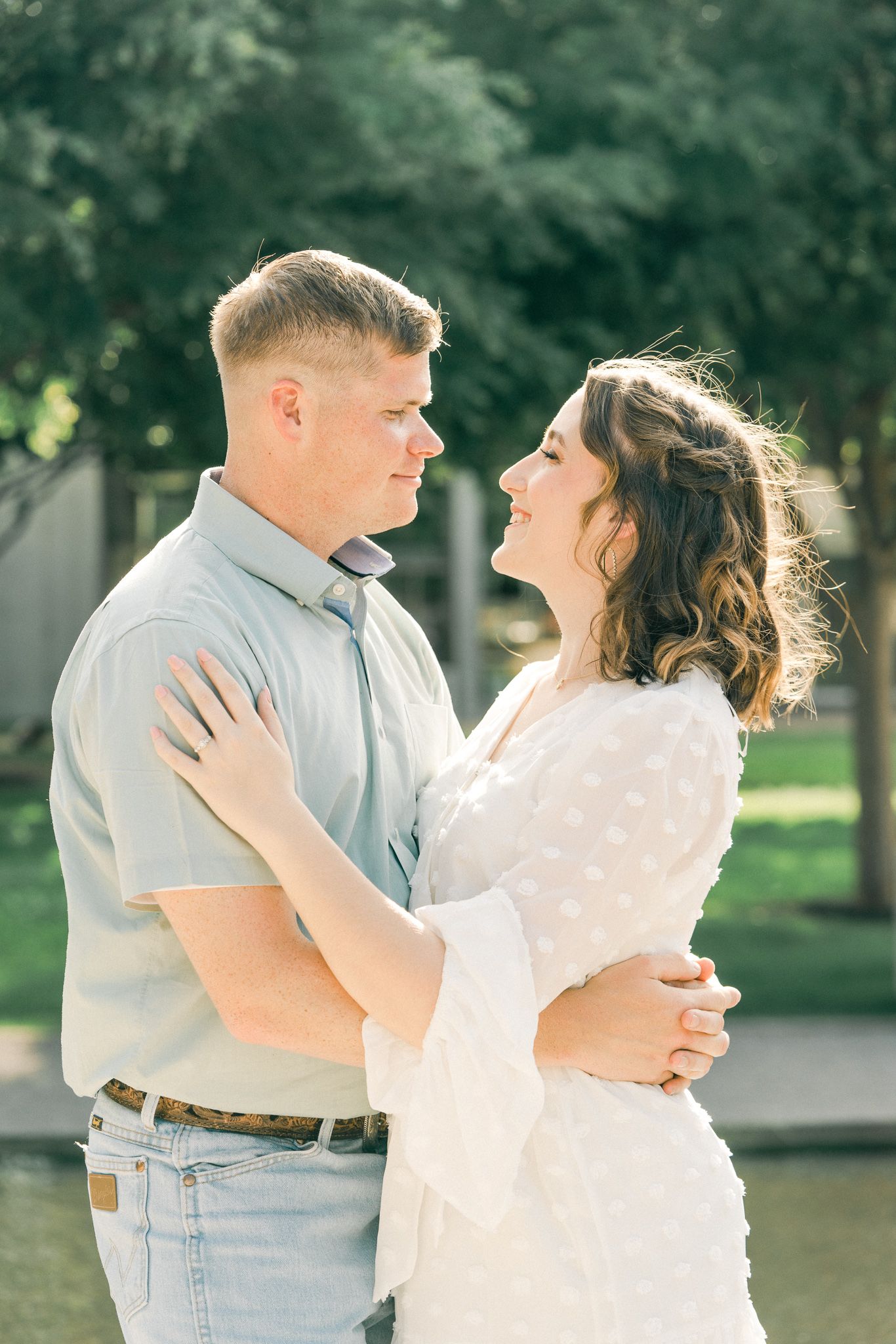 A romantic couple dances together outdoors in a park setting wearing white and light blue casual attire.