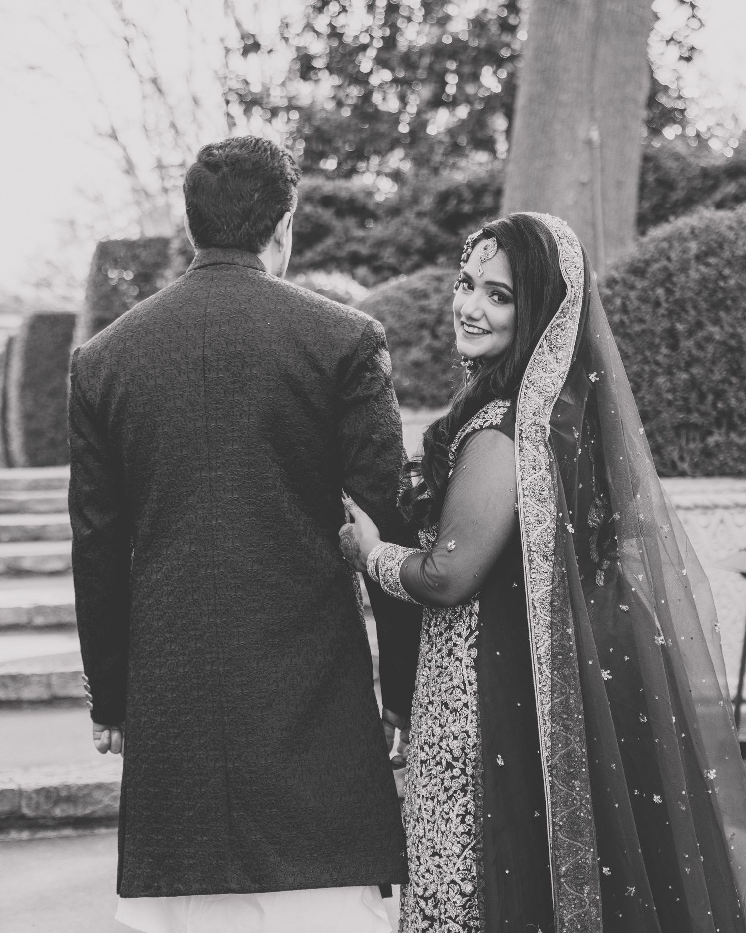 A black and white portrait of a couple in South Asian wedding attire sharing a romantic moment outdoors.