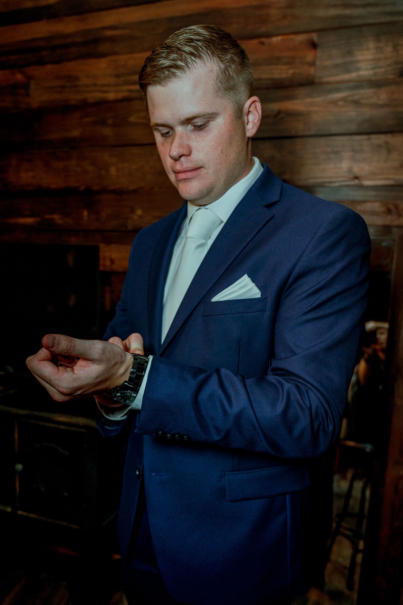 Someone wearing a navy blue suit adjusts their pocket square while standing against a rustic wooden wall.