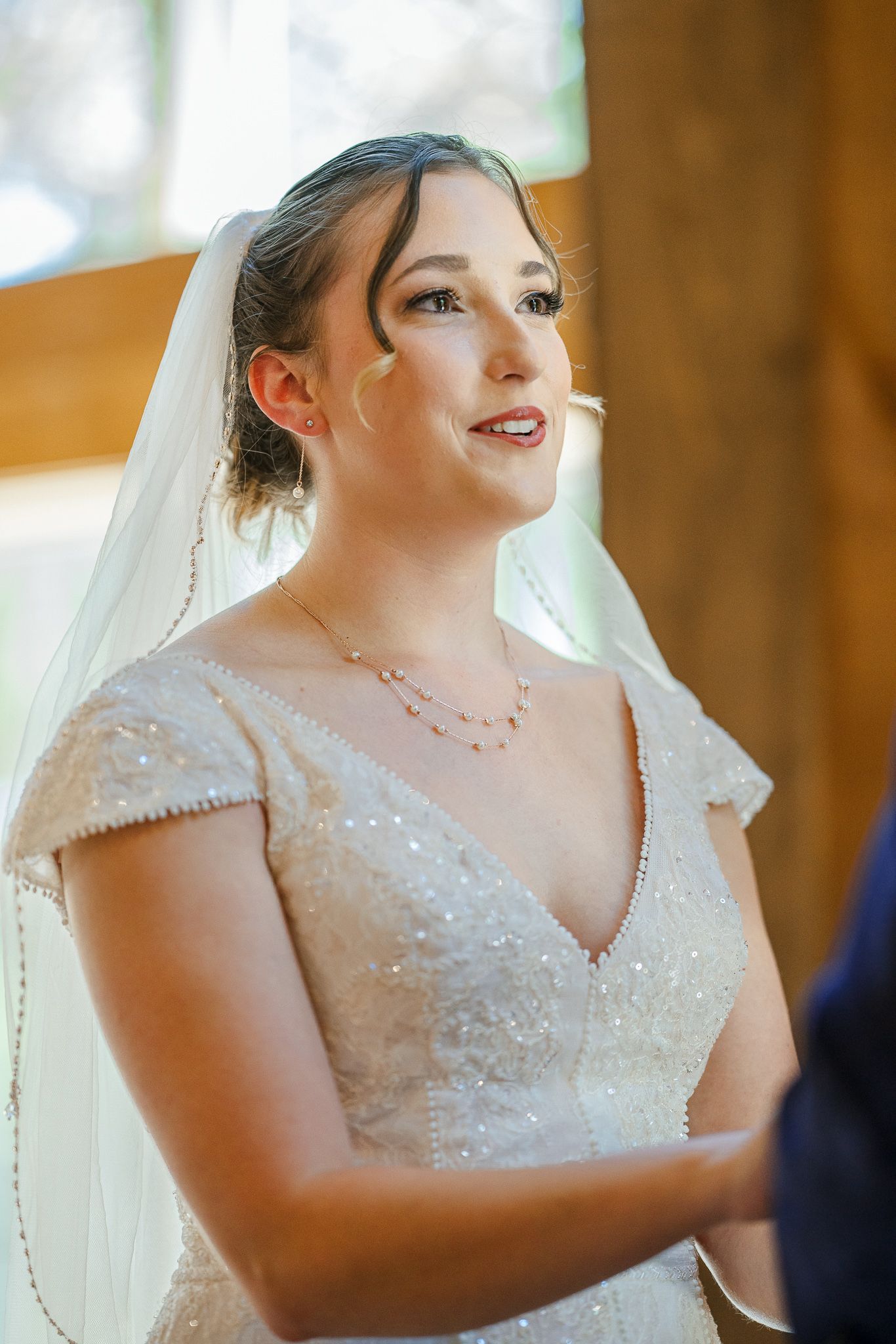 A bride in a sparkling white wedding dress and veil smiles joyfully during her wedding ceremony.