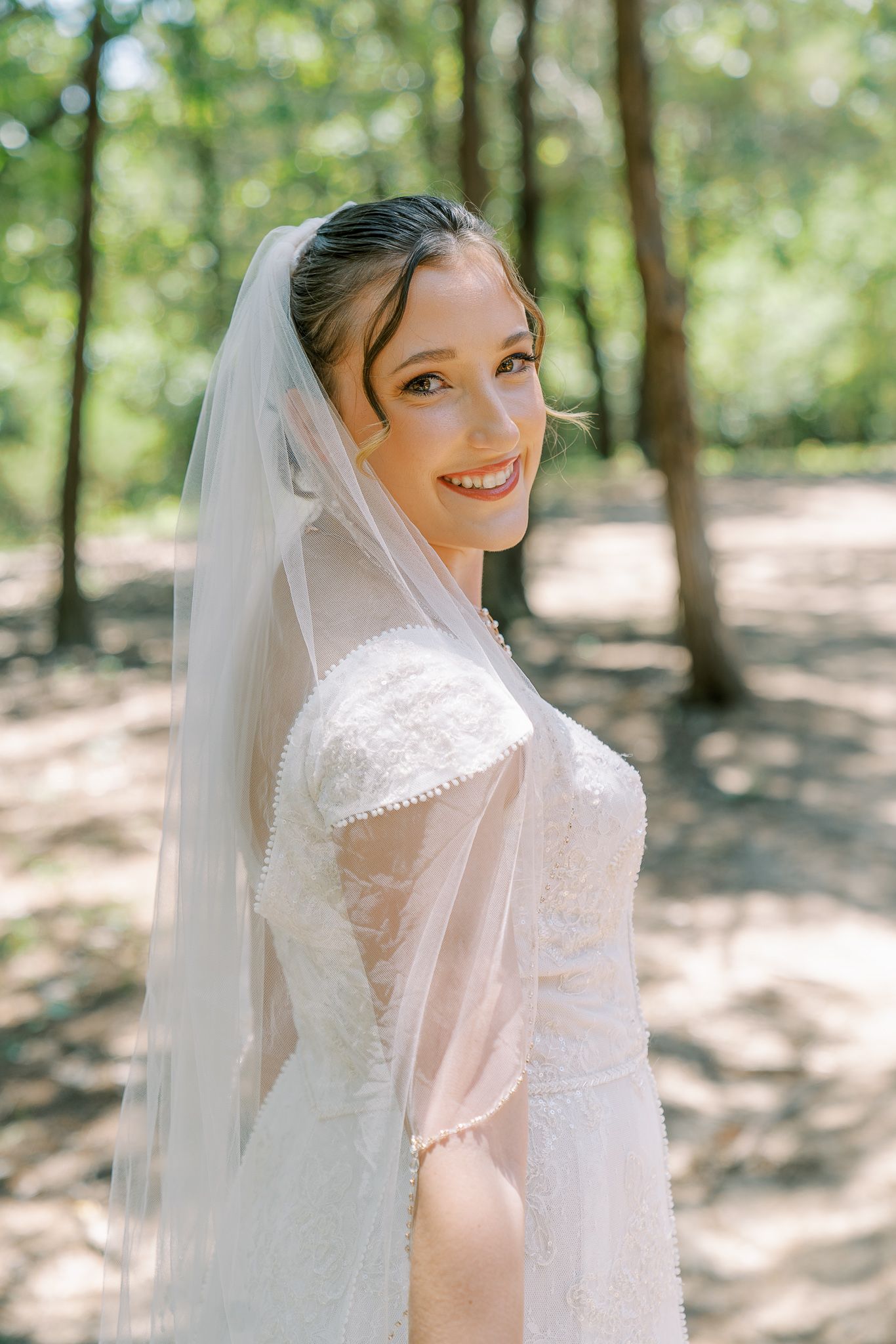 A bride in a white lace dress and flowing veil poses in a sunlit wooded area for romantic bridal portraits.