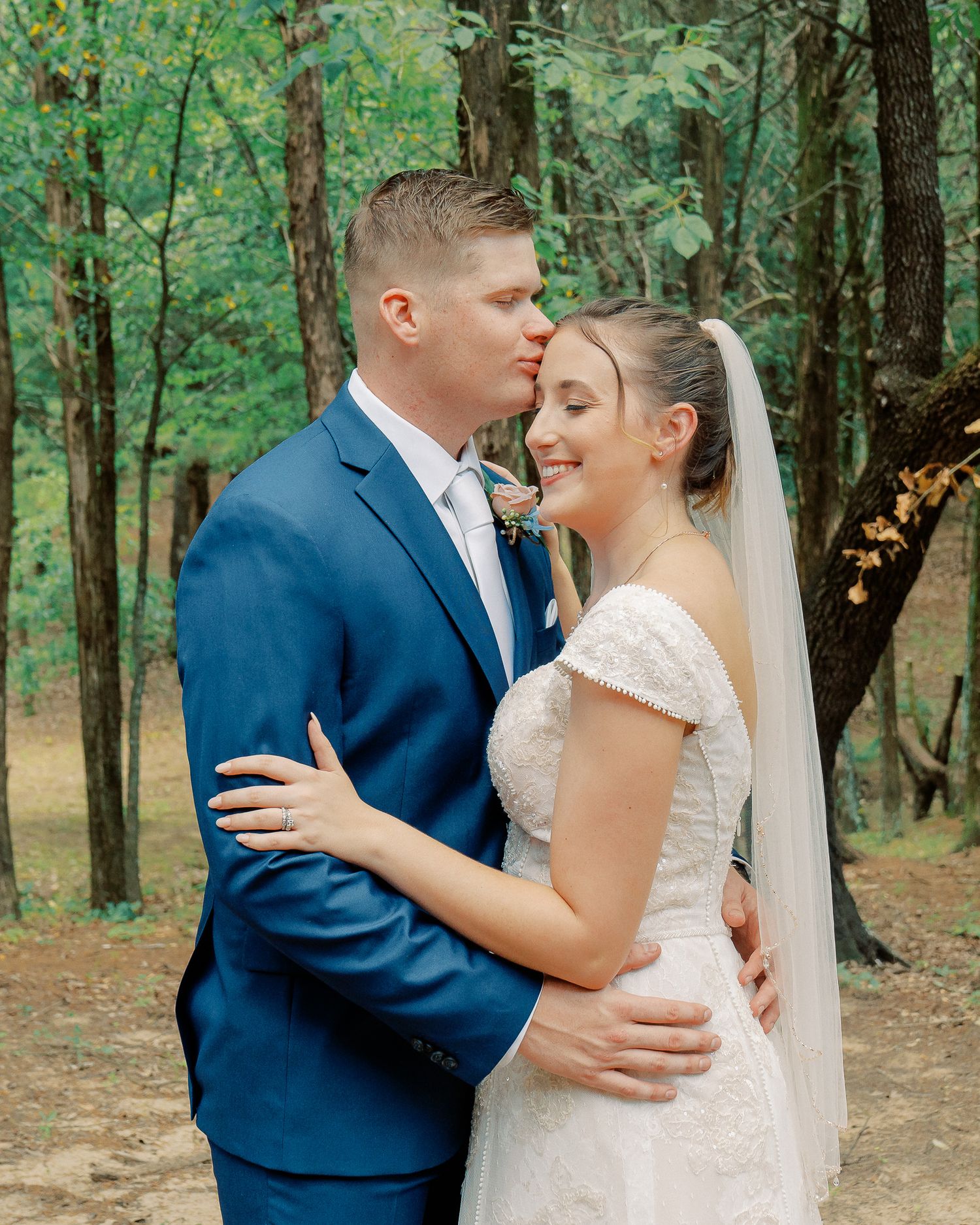Romantic wedding couple embraces in an intimate moment surrounded by forest trees and natural lighting.