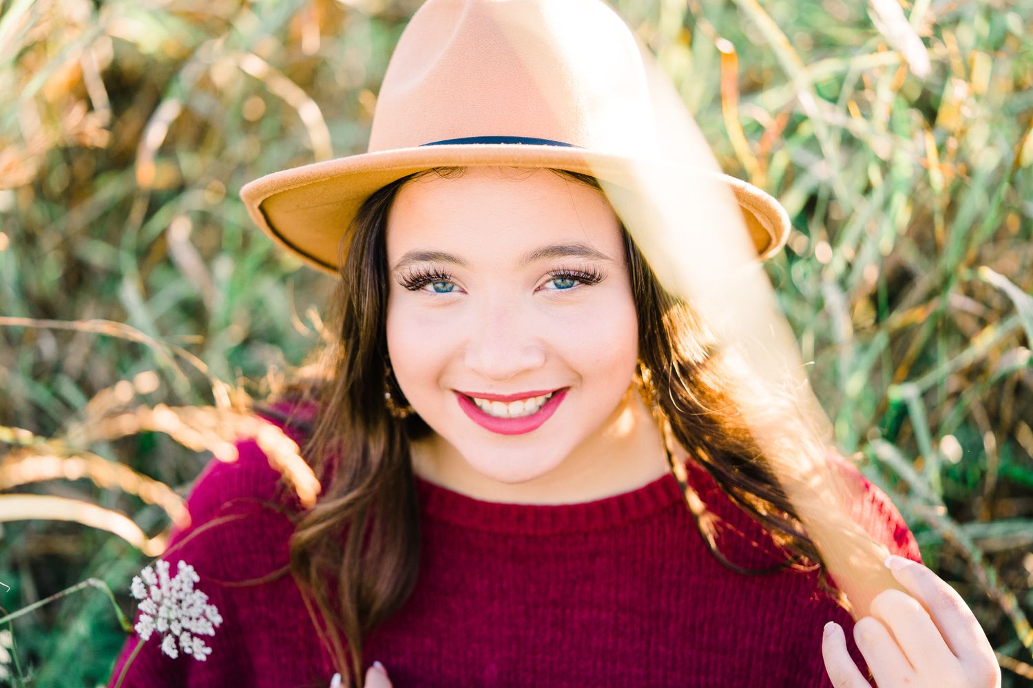 A person wearing a burgundy sweater and tan hat smiles brightly against a natural outdoor background.