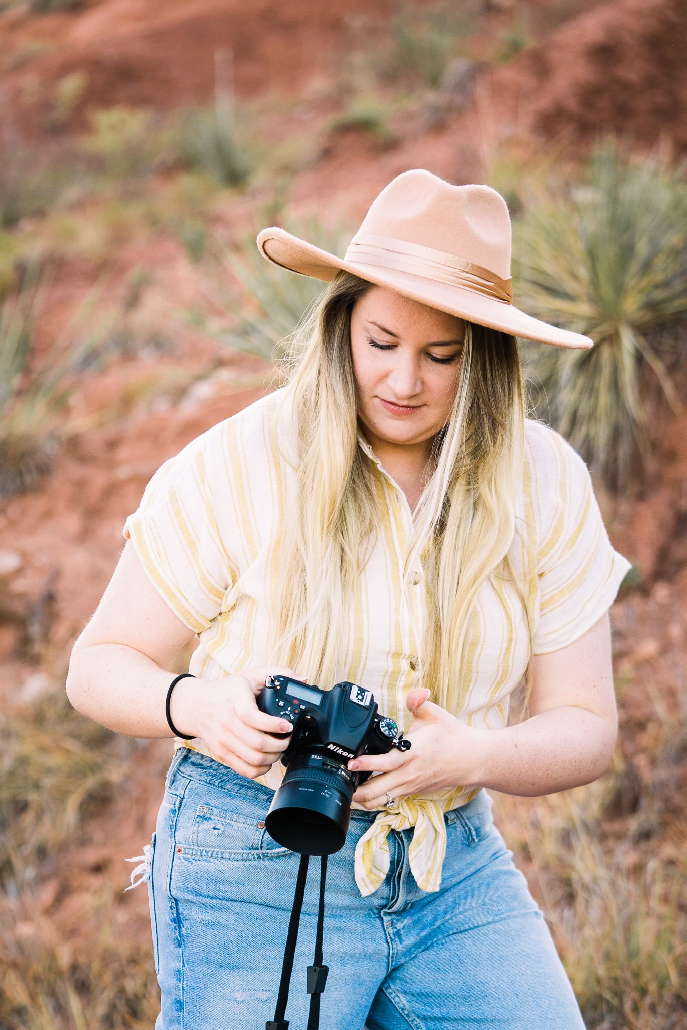 Photographer in a wide-brim hat and jeans adjusts camera settings while working outdoors in desert terrain.