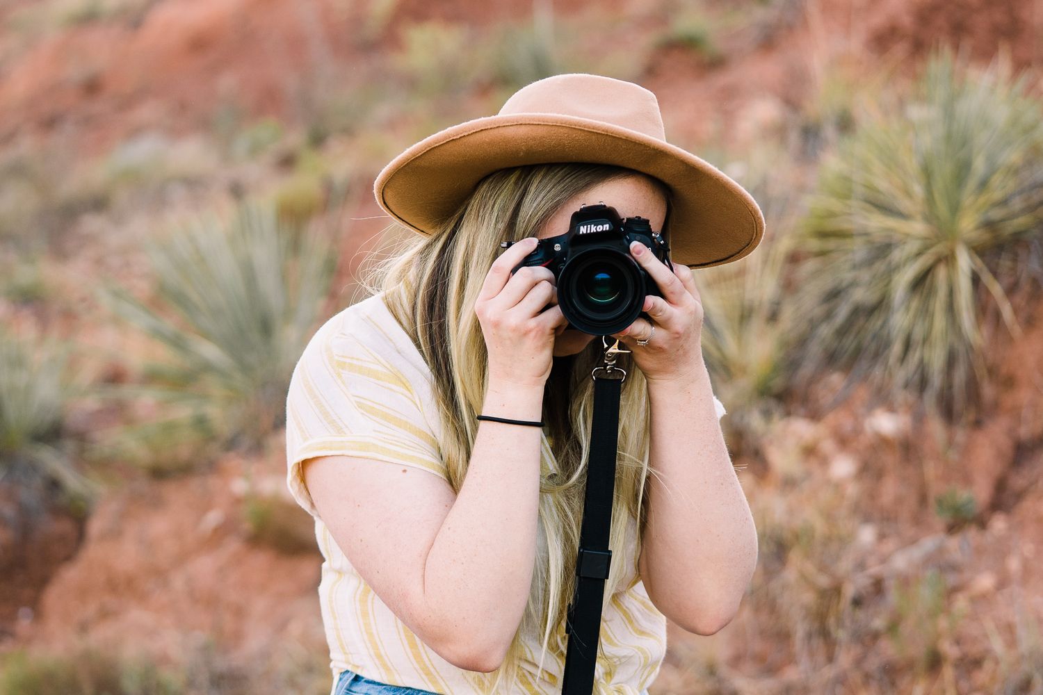 A photographer wearing a tan hat captures images in a desert landscape with red rocks and desert plants in the background.