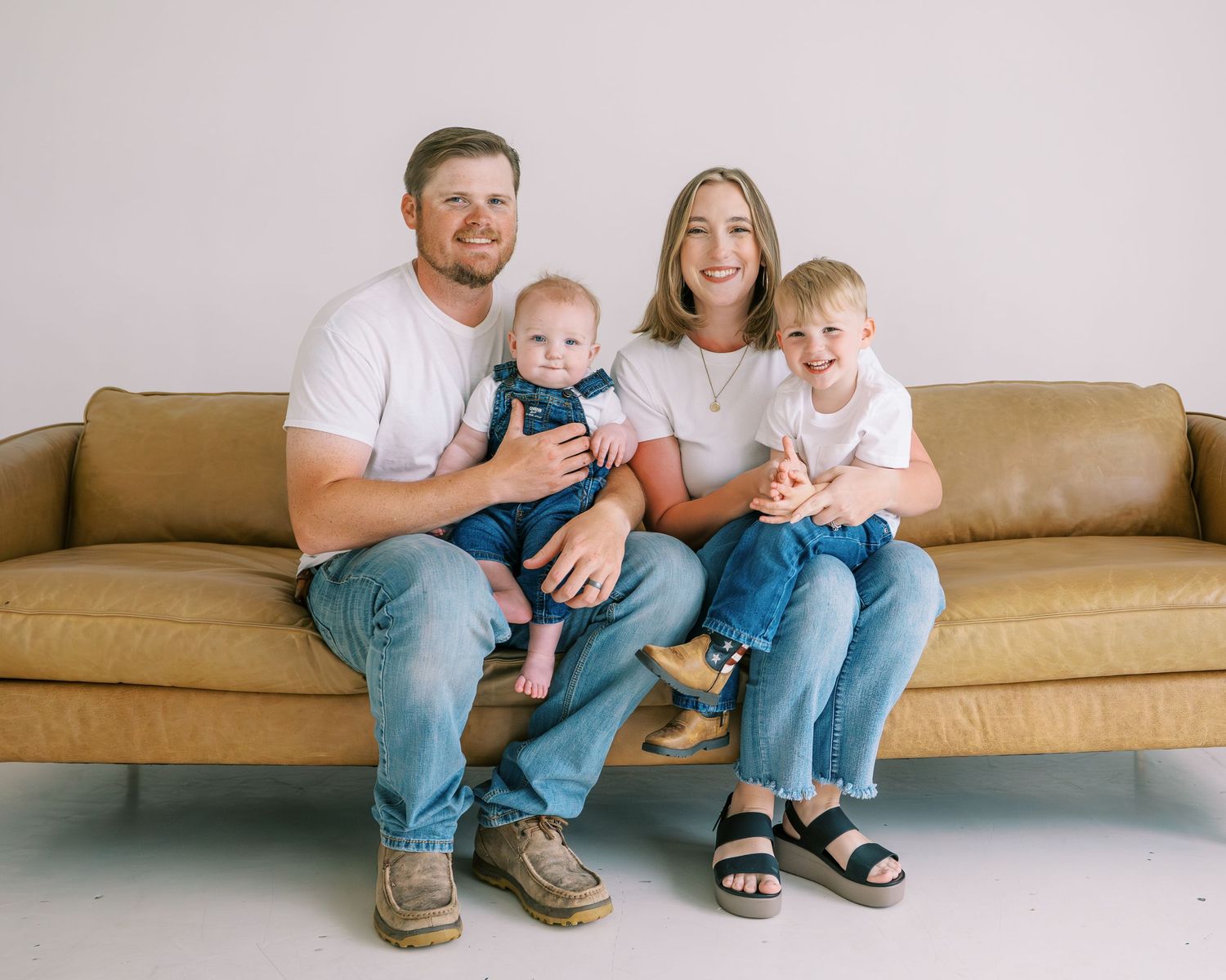 Happy family of four posing on a tan leather couch, parents in white tees and jeans with two young children.