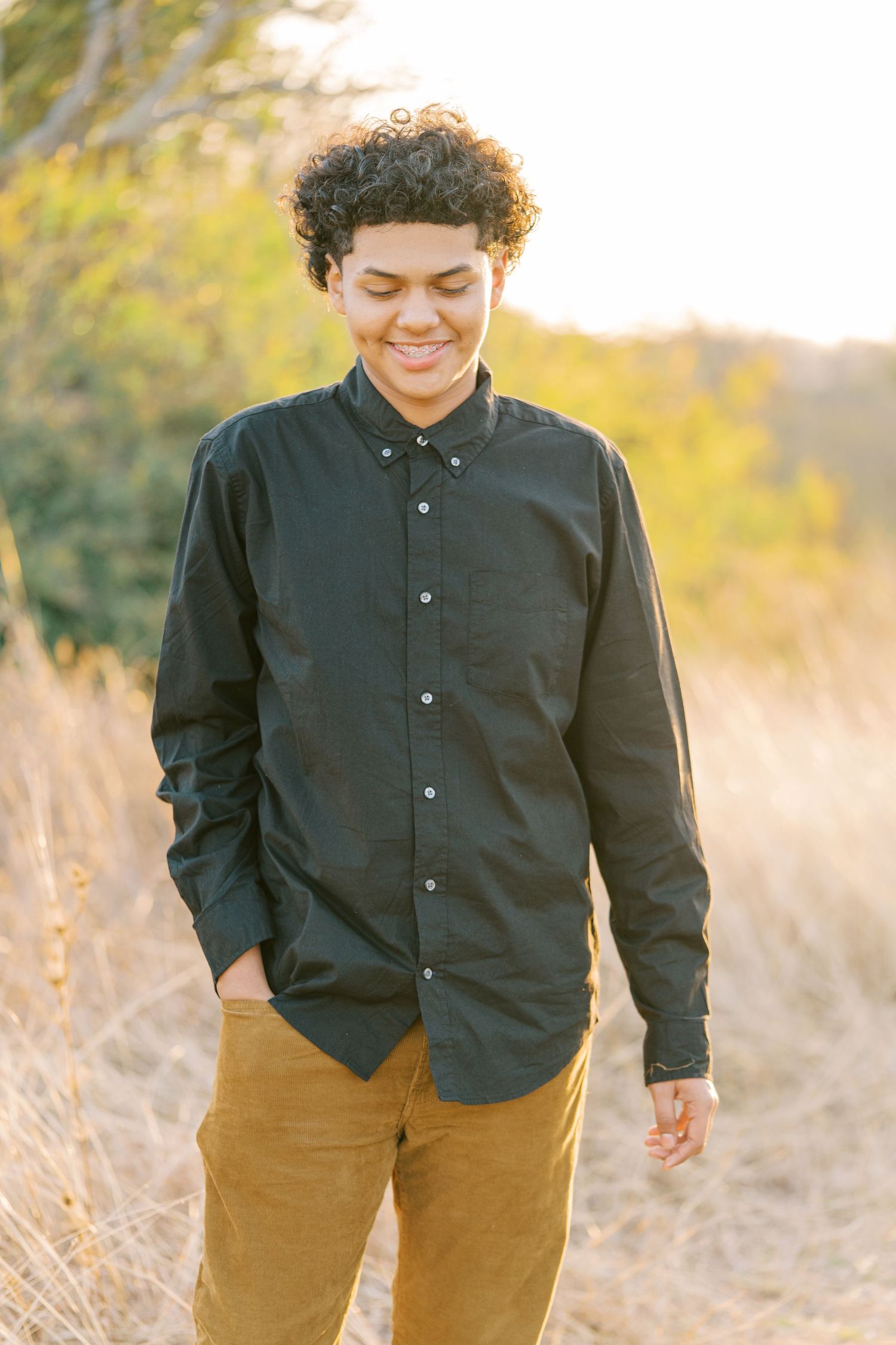Smiling young person with curly hair wearing a black shirt and tan pants standing in a sunlit field.