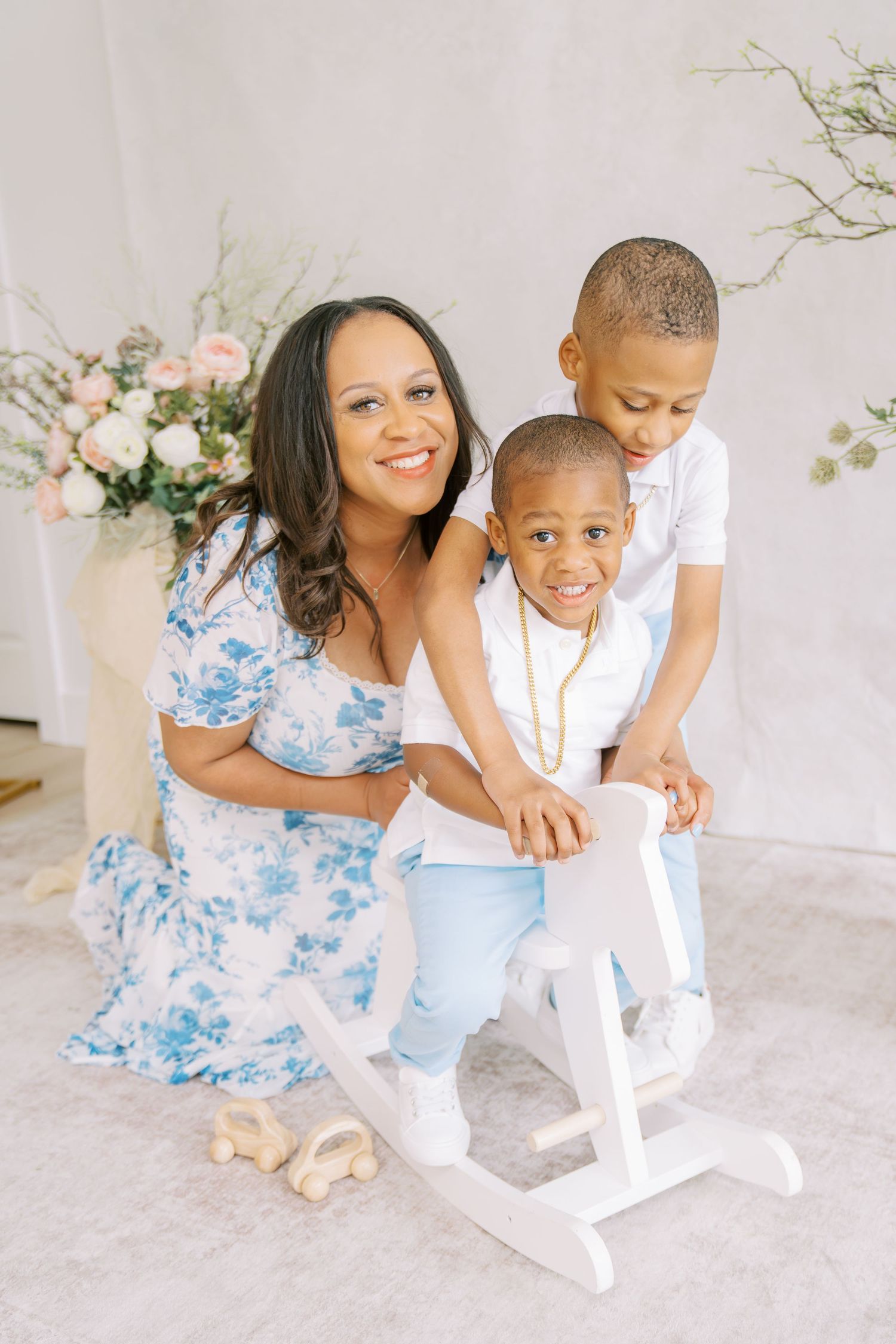 A smiling woman in a floral dress poses with two young boys near a white rocking horse in a bright studio setting.