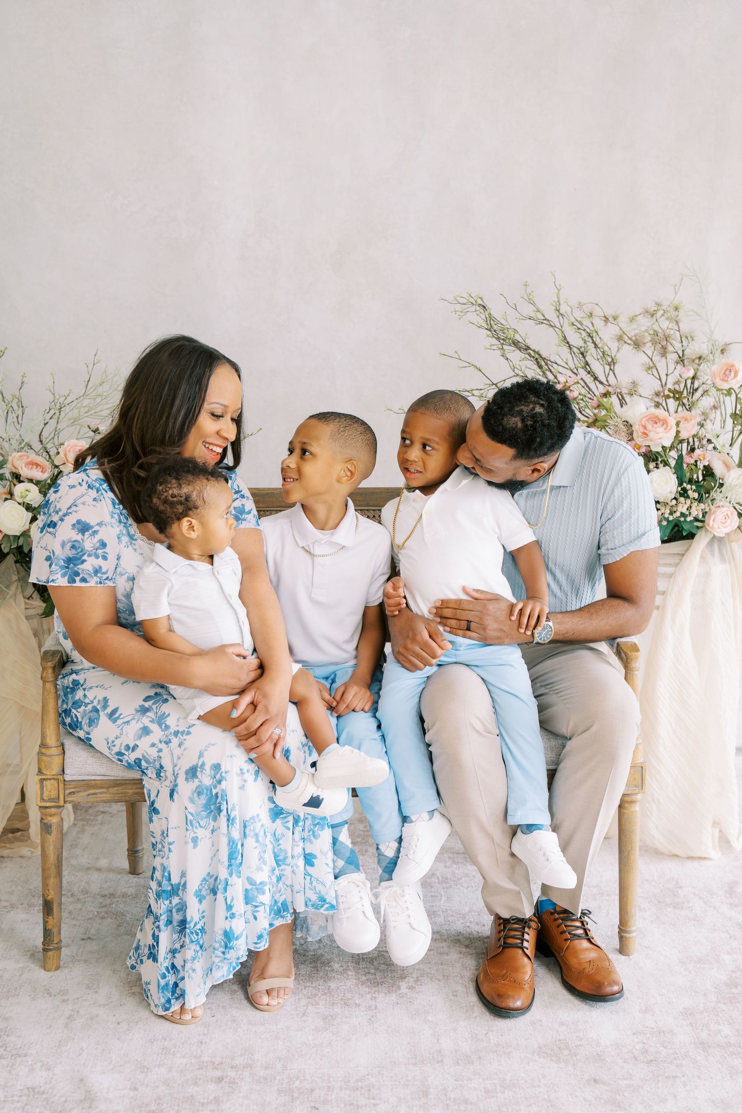A loving Black family of four poses together on a bench with floral arrangements in a bright studio portrait.