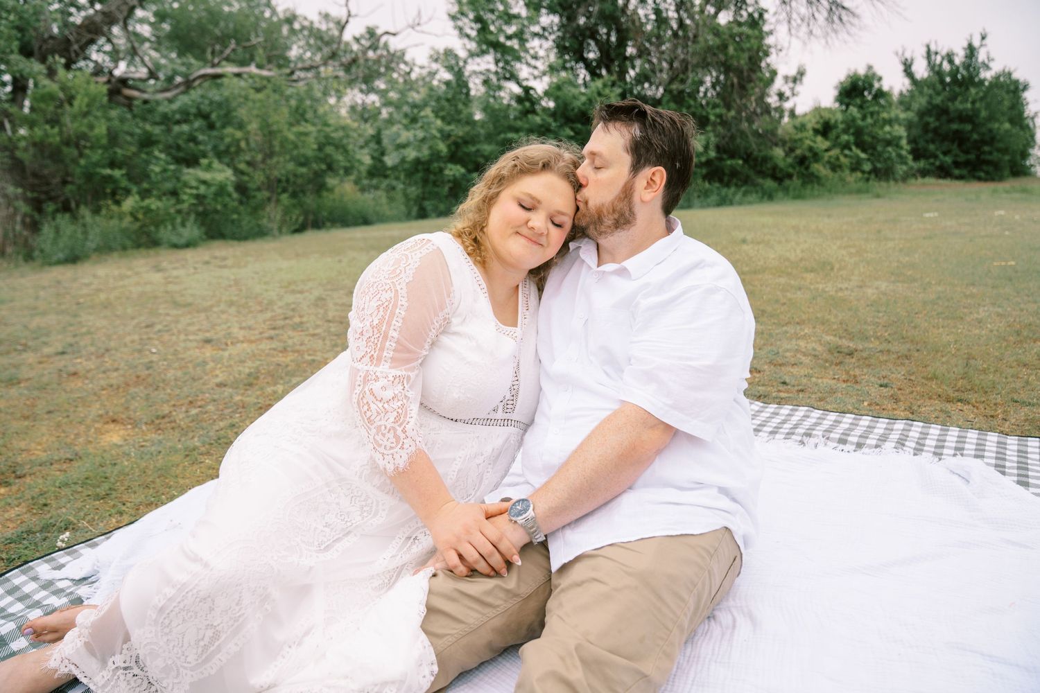 Couple sitting on a picnic blanket outdoors, man kissing woman on cheek as she leans into him smiling.