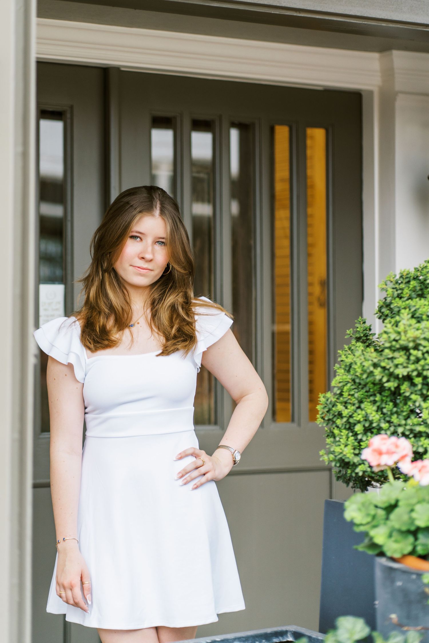 A person in a white summer dress stands on a porch with potted plants and greenery in the background.