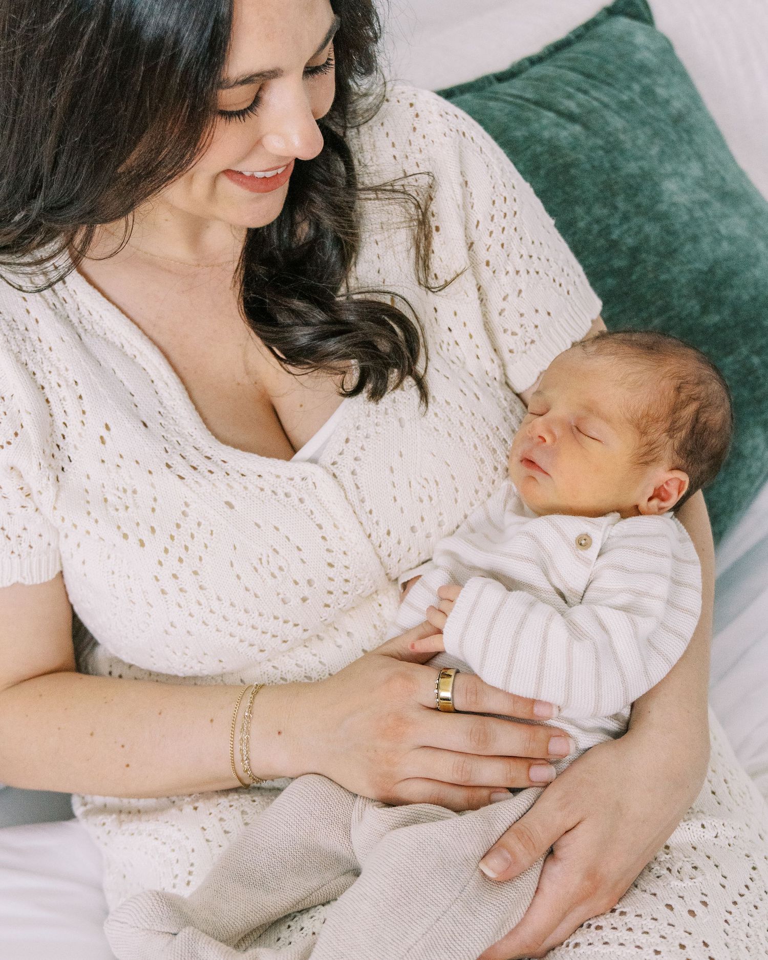Parent in white eyelet dress cradles sleeping newborn baby on emerald green velvet couch in tender moment.