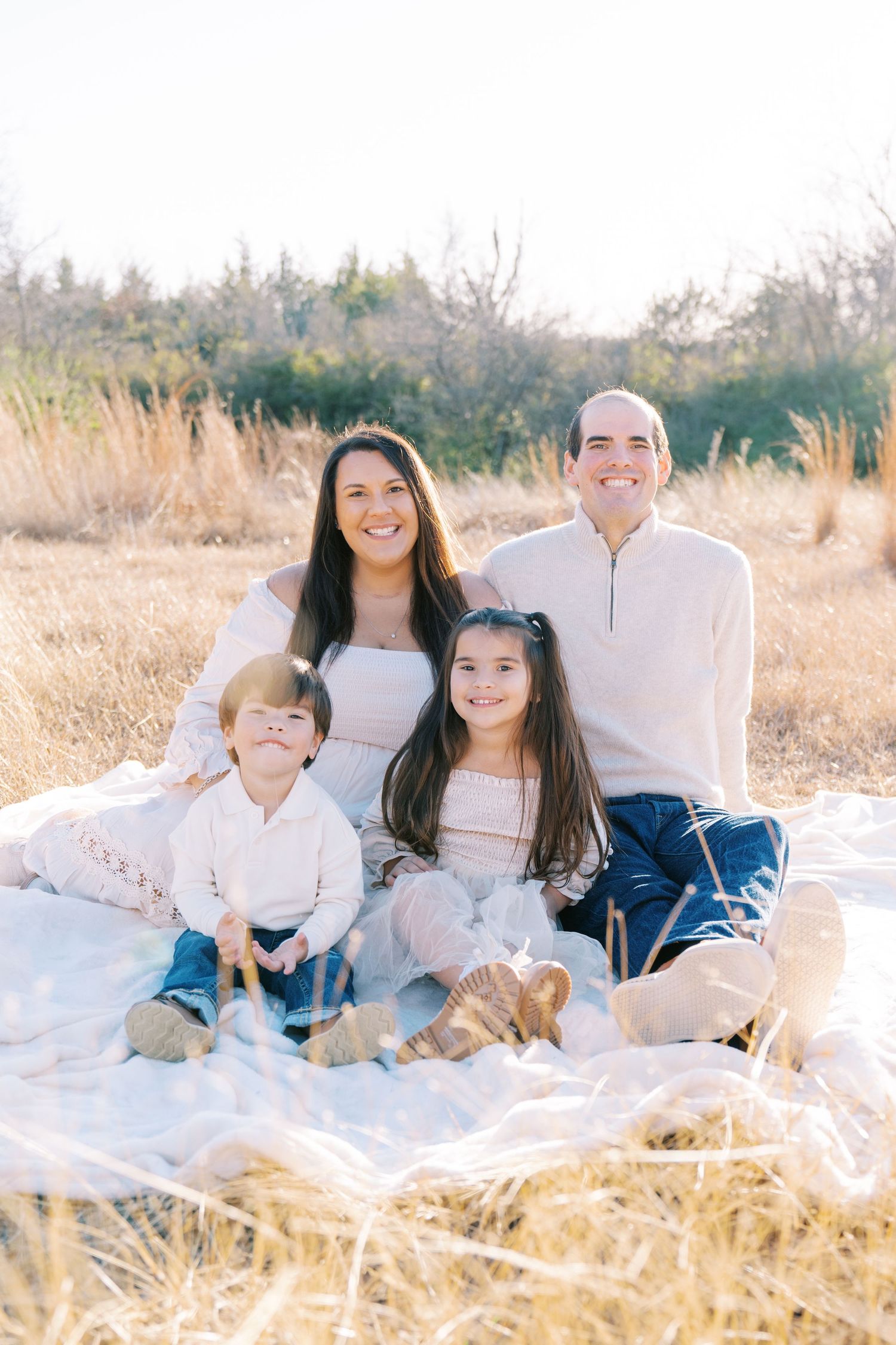 Family sits together on white blanket in field during outdoor photo session.